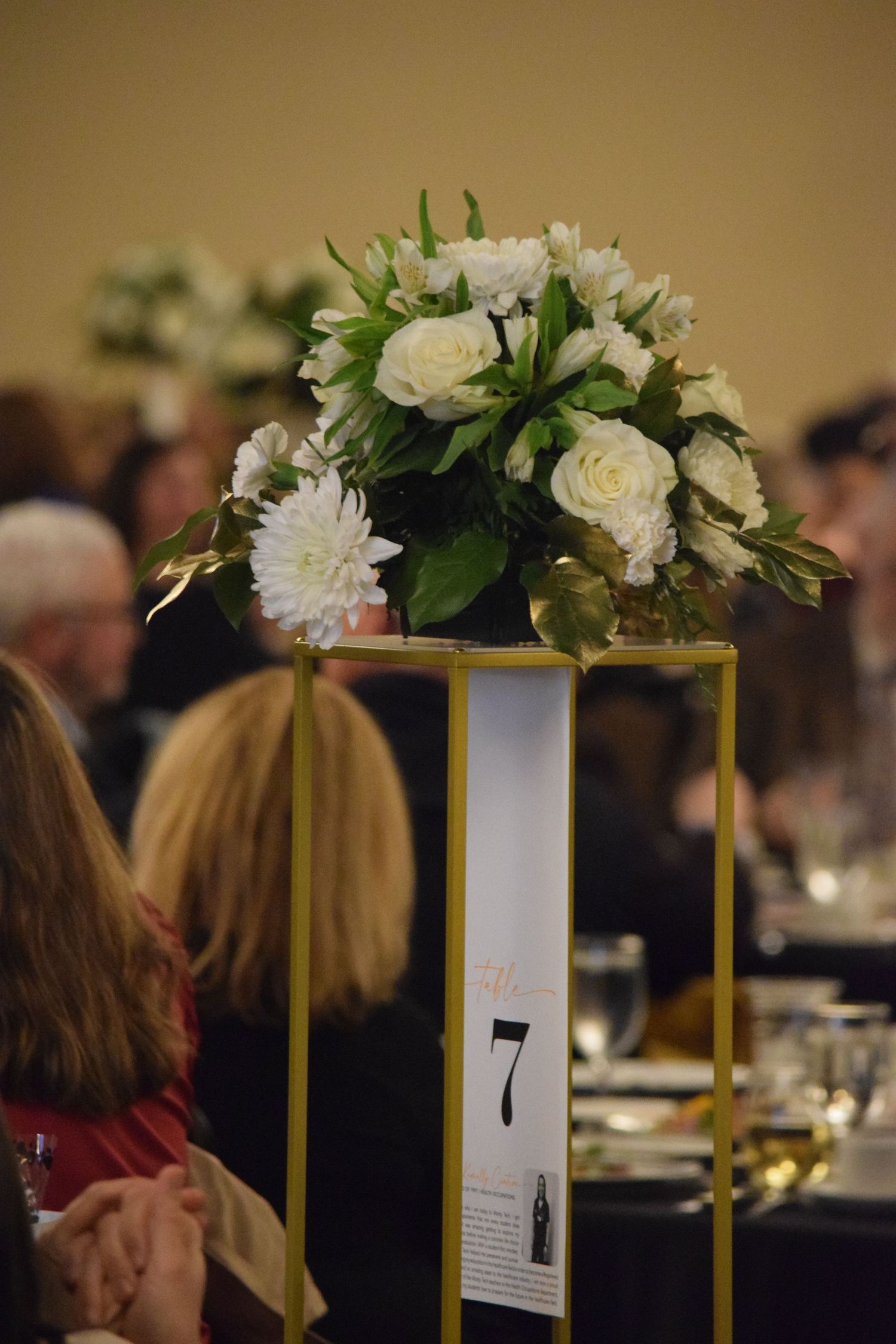 Elegant white rose and chrysanthemum centerpiece in gold geometric stand with table number 7 at formal event.