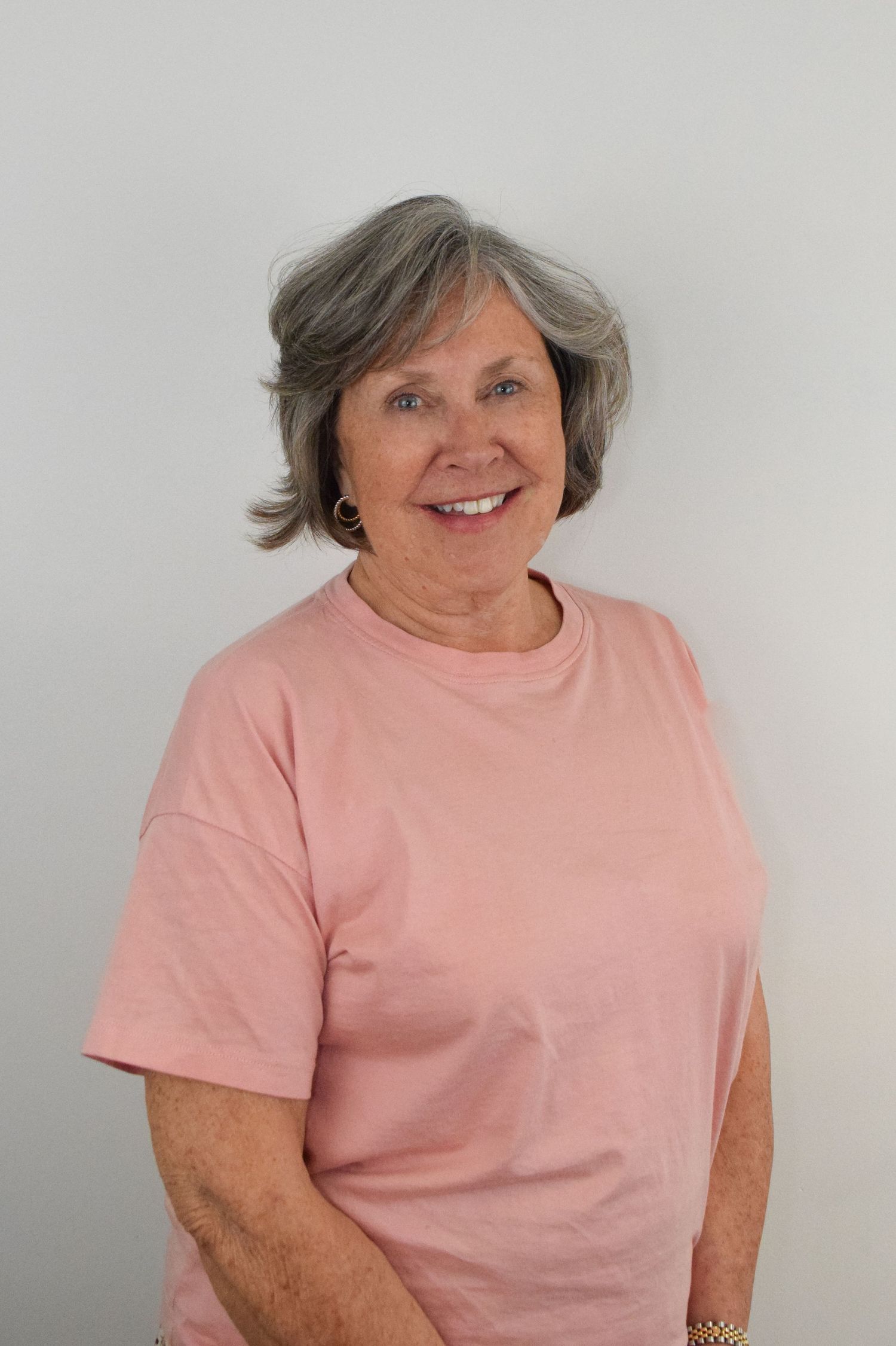 An older woman wearing a pink t-shirt stands against a light gray background, smiling warmly at the camera.