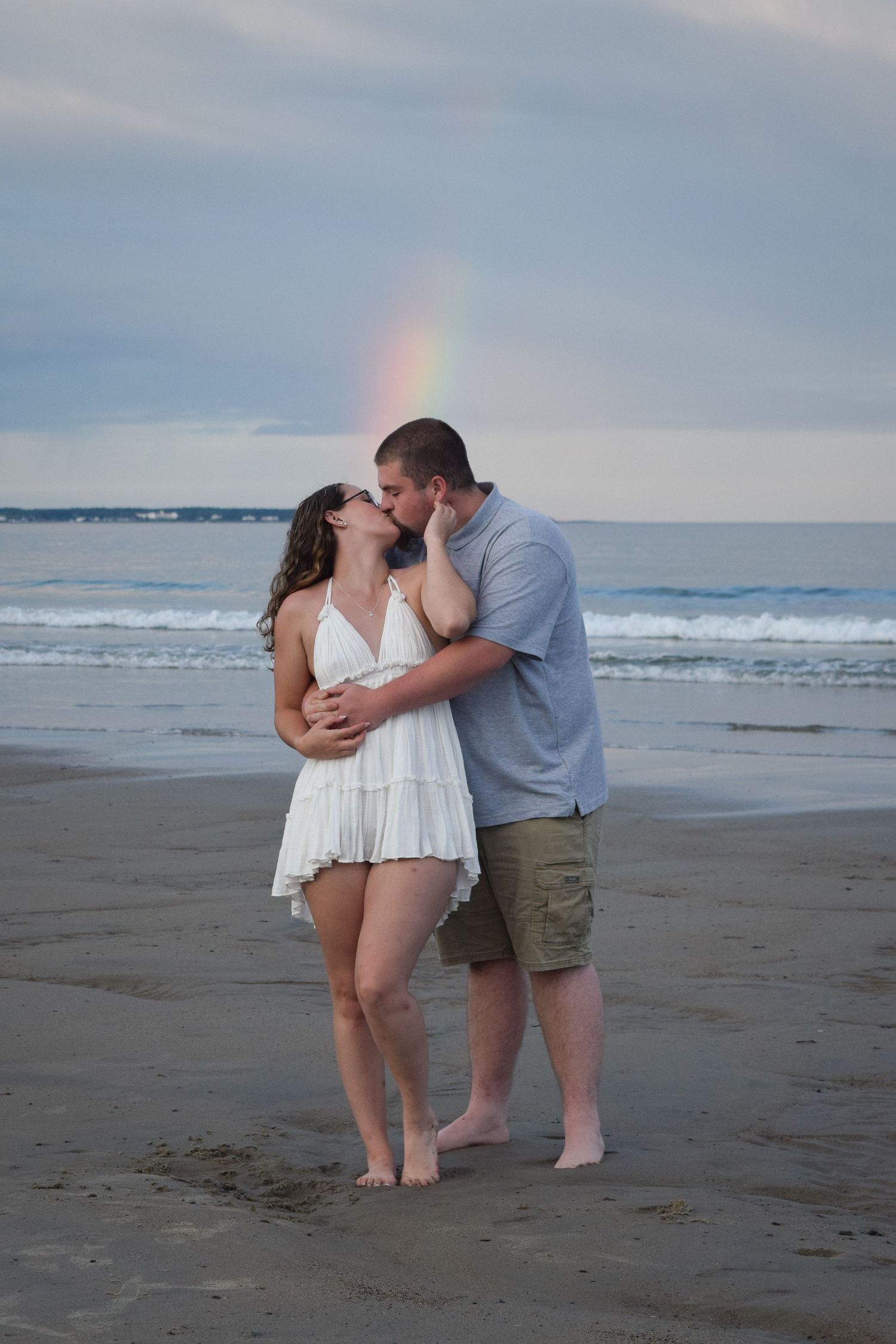 A romantic couple shares tender moments on a cloudy beach with ocean waves and a rainbow in the background.