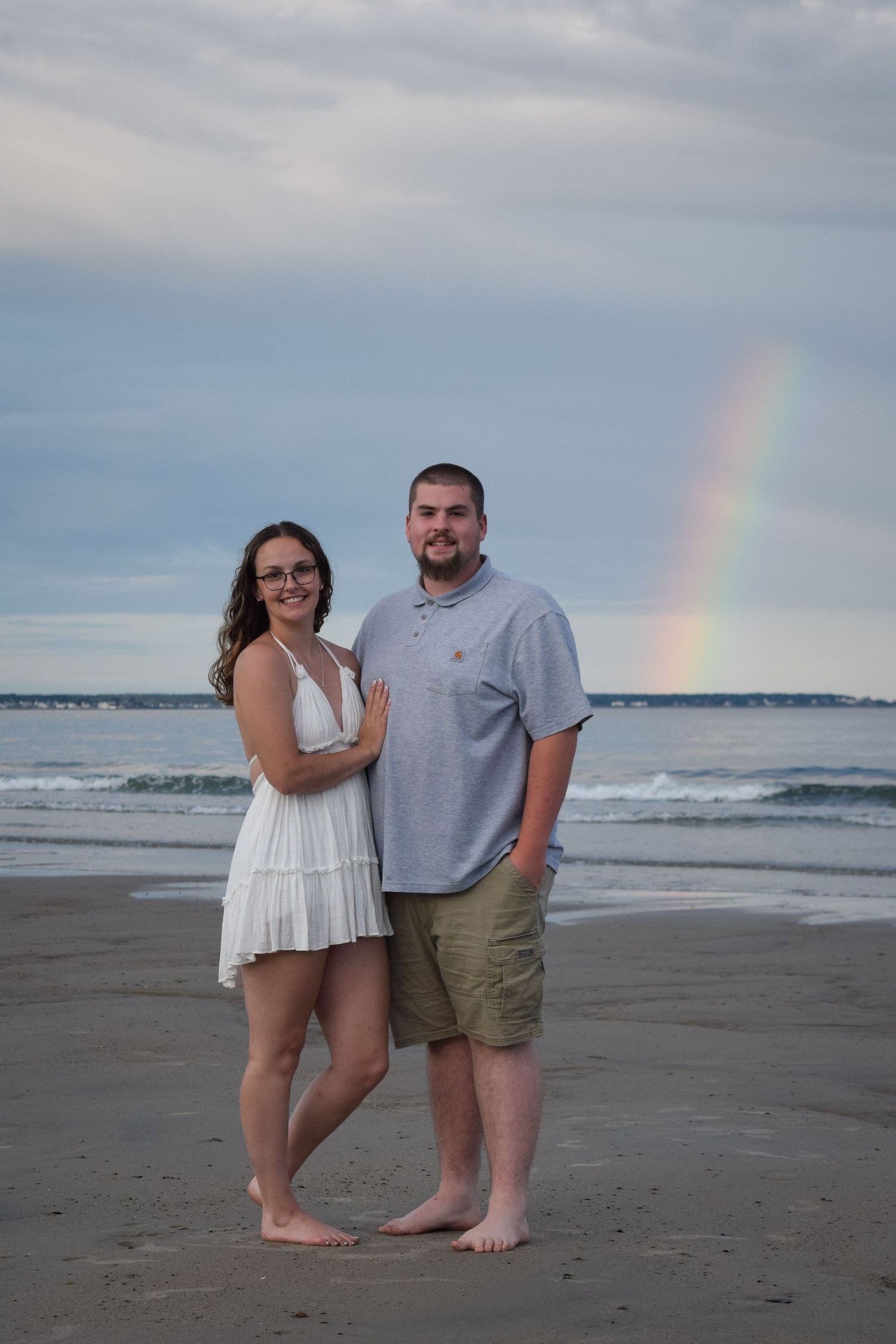 A couple stands barefoot on the beach at sunset with a rainbow appearing in the cloudy sky over the ocean.