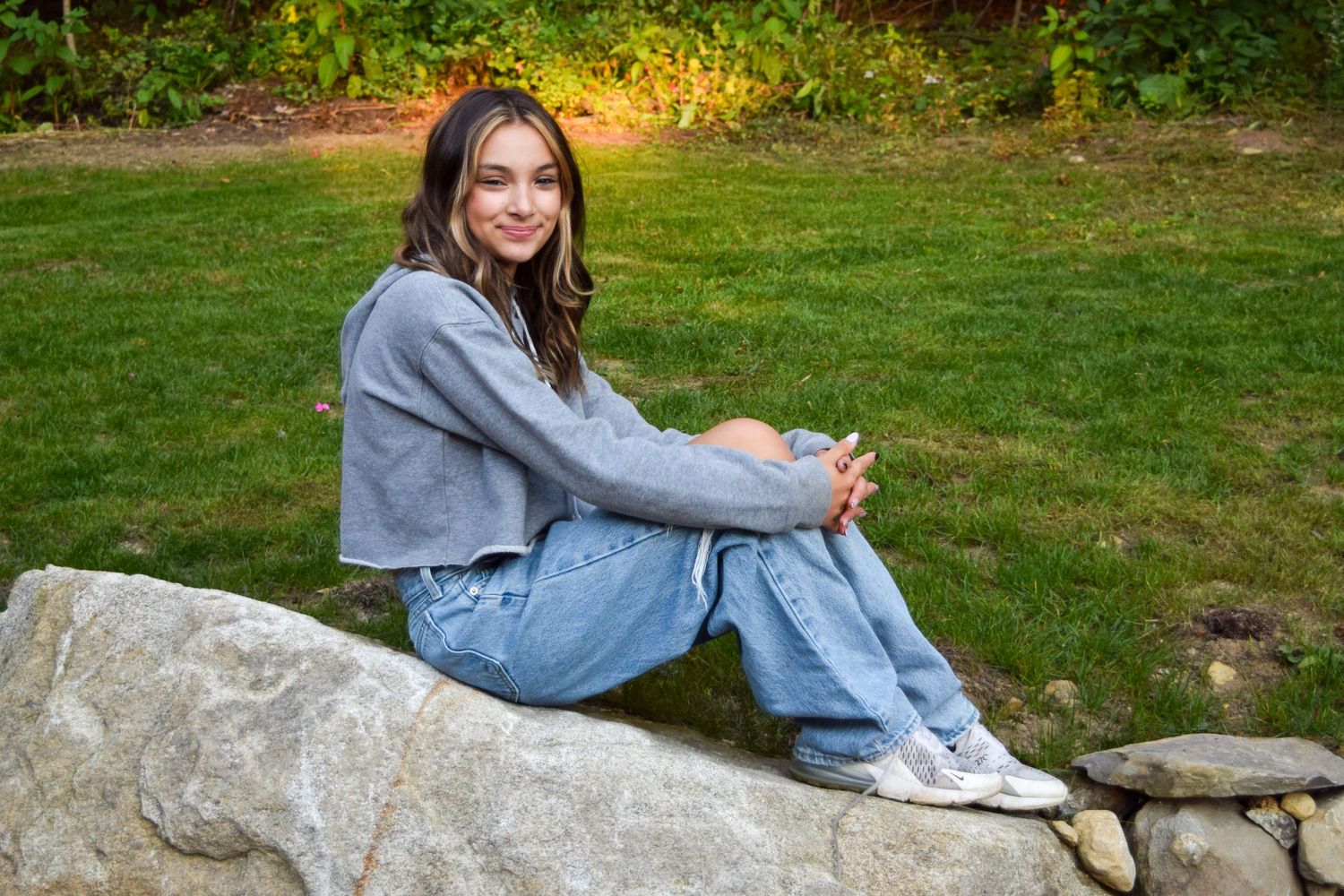 A person in casual denim clothing sits on a stone wall in a grassy outdoor setting during a sunny day.