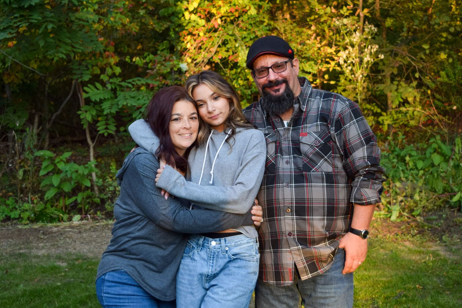 A family of three poses together outdoors in casual clothing with autumn foliage in the background.