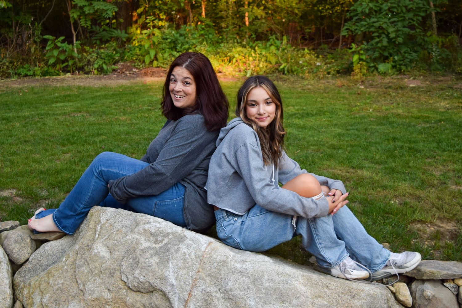 Two people sit back-to-back on a large rock, wearing casual denim and sweatshirts in an outdoor setting with green grass.