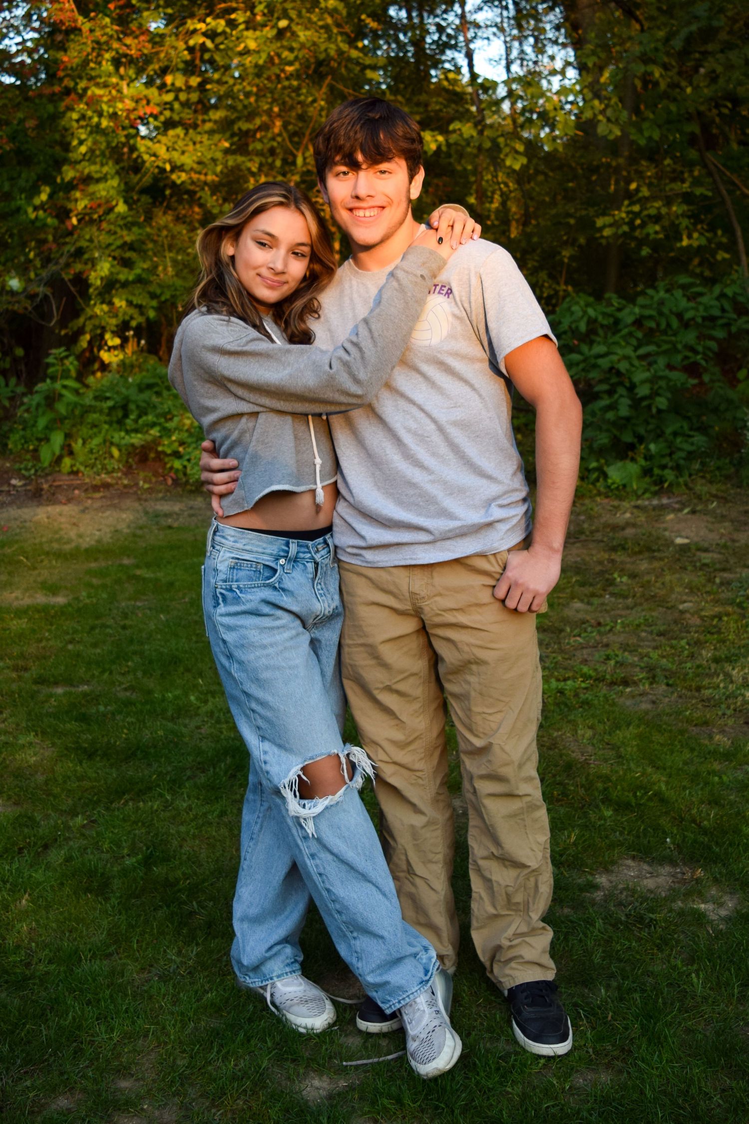 Two people wearing casual outfits embrace outdoors in a natural setting with autumn foliage in the background.