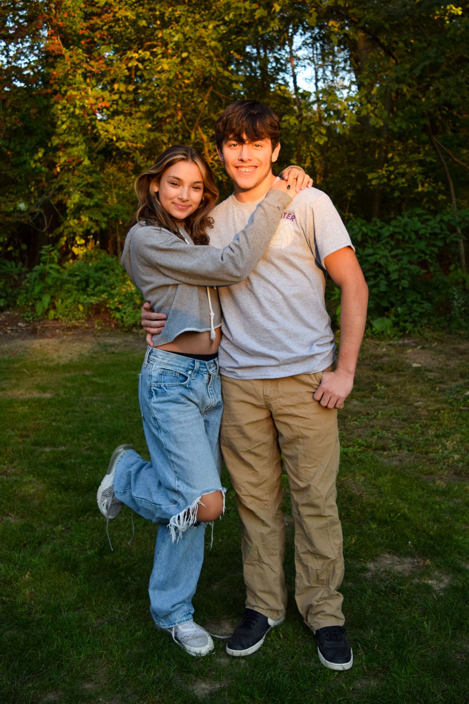 Two young people pose together outdoors in casual clothing against an autumn forest backdrop during golden hour.