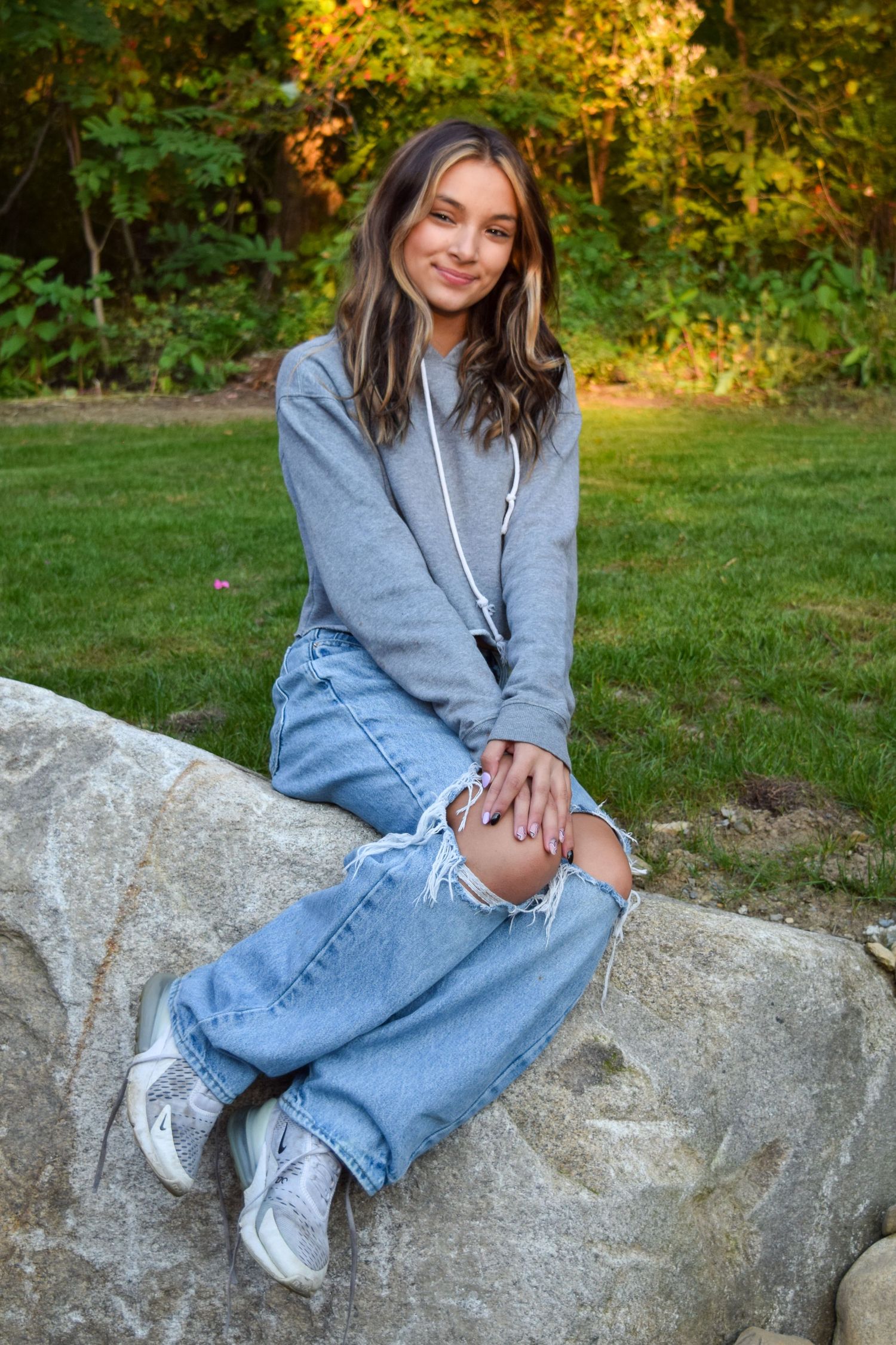 A person in a gray hoodie and jeans sits casually on a large rock outdoors during golden hour.