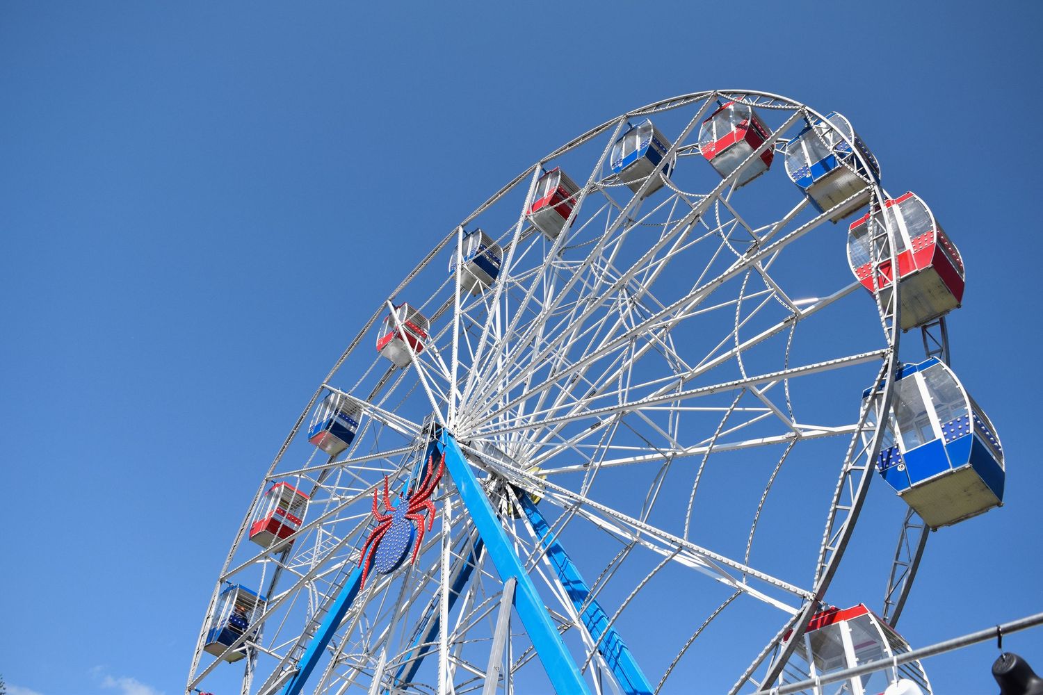 A Ferris wheel with red and white gondolas stands against a bright blue sky at a carnival or fairground.