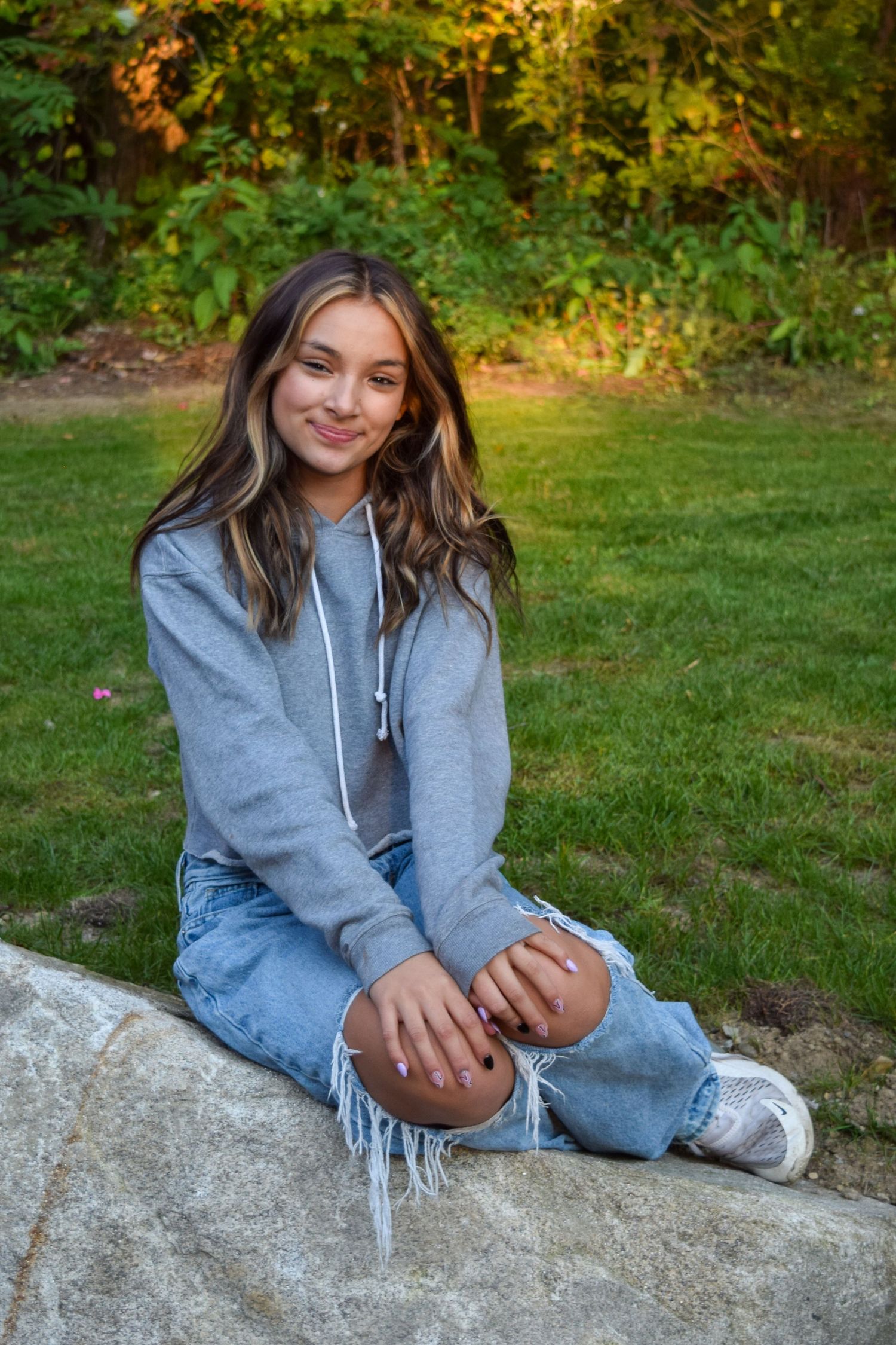 Someone in casual attire with a grey hoodie and ripped jeans sits on a large rock at sunset in a grassy outdoor setting.