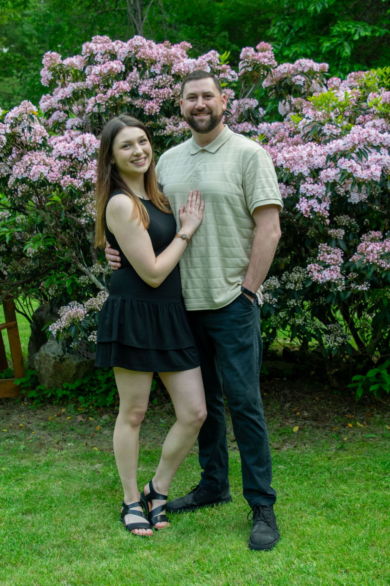 A couple poses together in front of blooming pink rhododendron bushes in a lush garden setting.