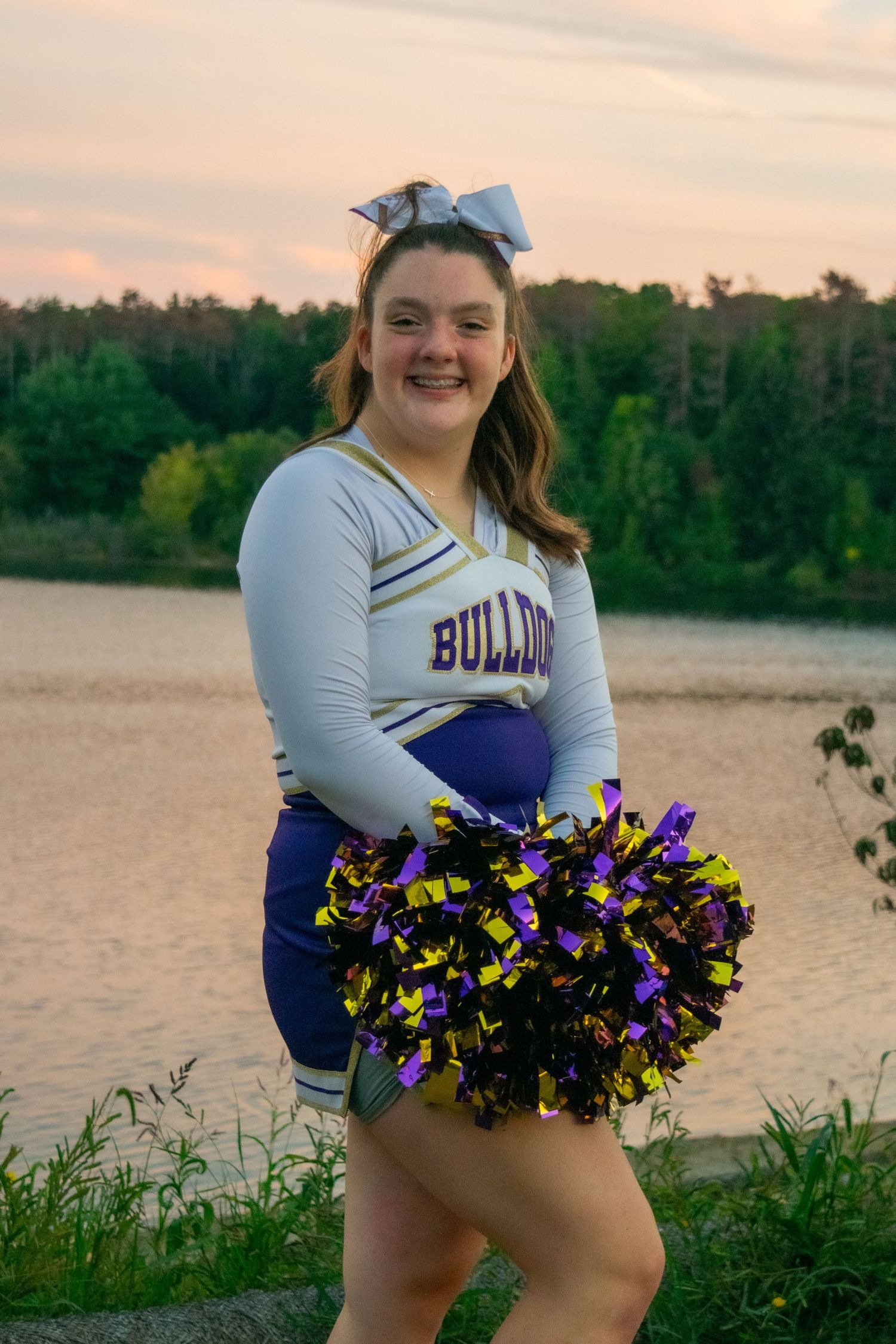 A cheerleader in a blue and white uniform holds purple and gold pom poms while posing outdoors at sunset.