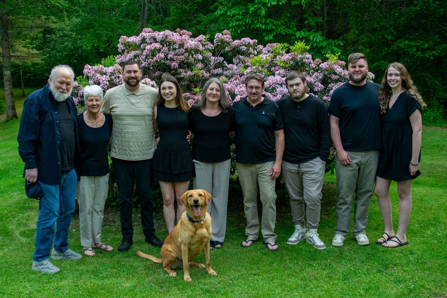 A large group of people and a golden retriever dog gather together for an outdoor photo in a green lawn setting.