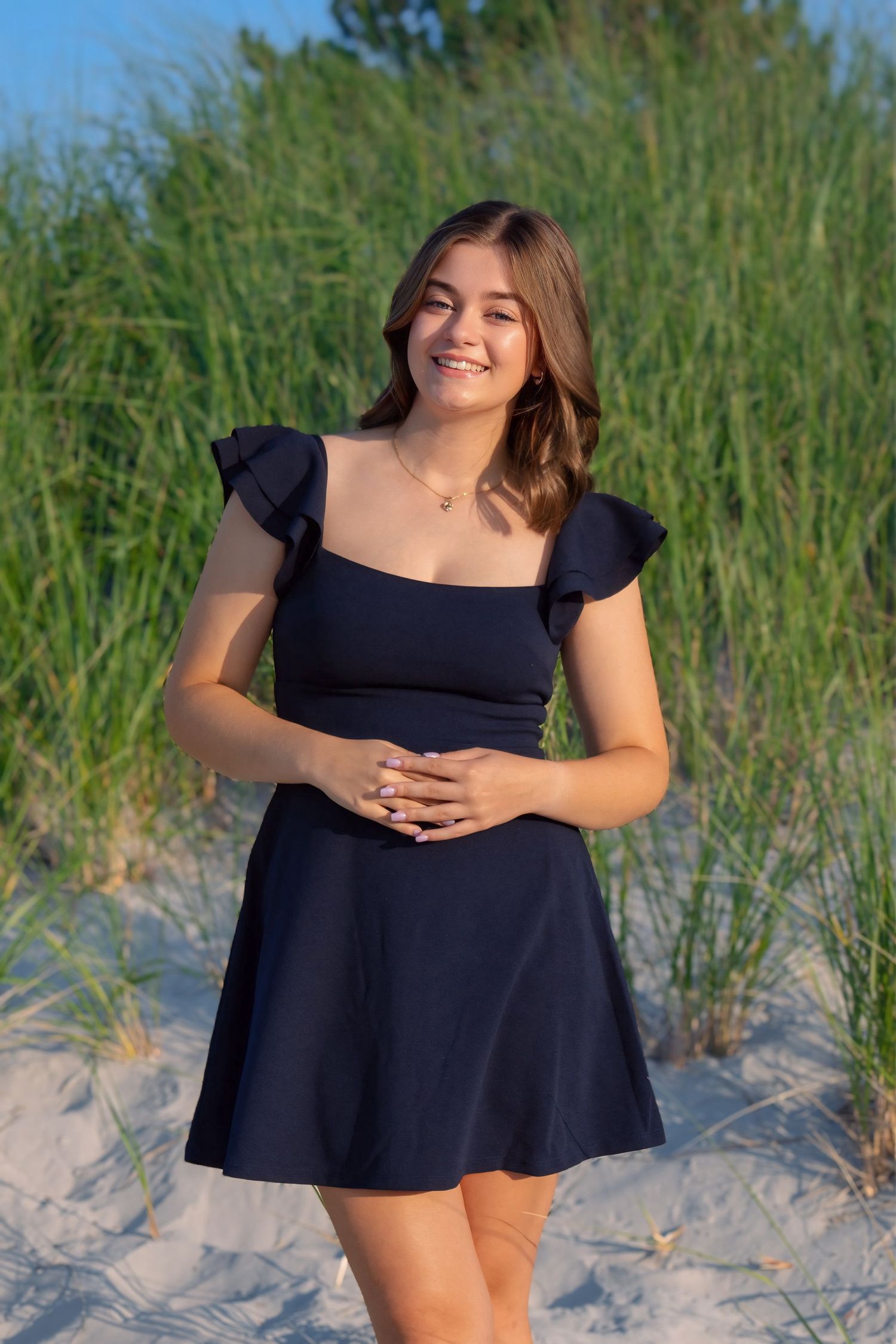 A person in a navy blue summer dress poses on a sandy beach with tall grass in the background during golden hour.