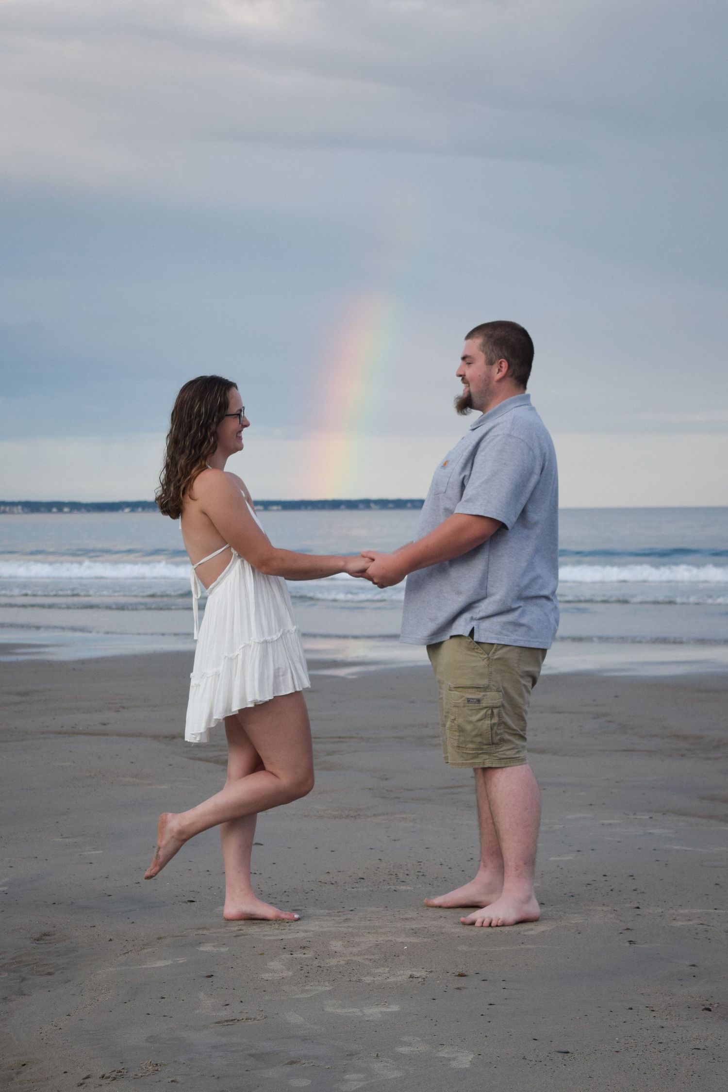 A couple stands on a beach at sunset with a rainbow in the background as they playfully pose for photos.