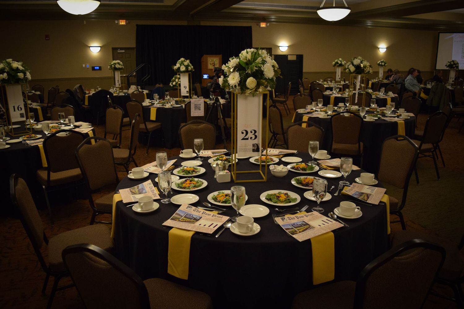 An elegant banquet table setup with navy blue linens, yellow accents, and white floral centerpiece in a dimly lit reception hall.