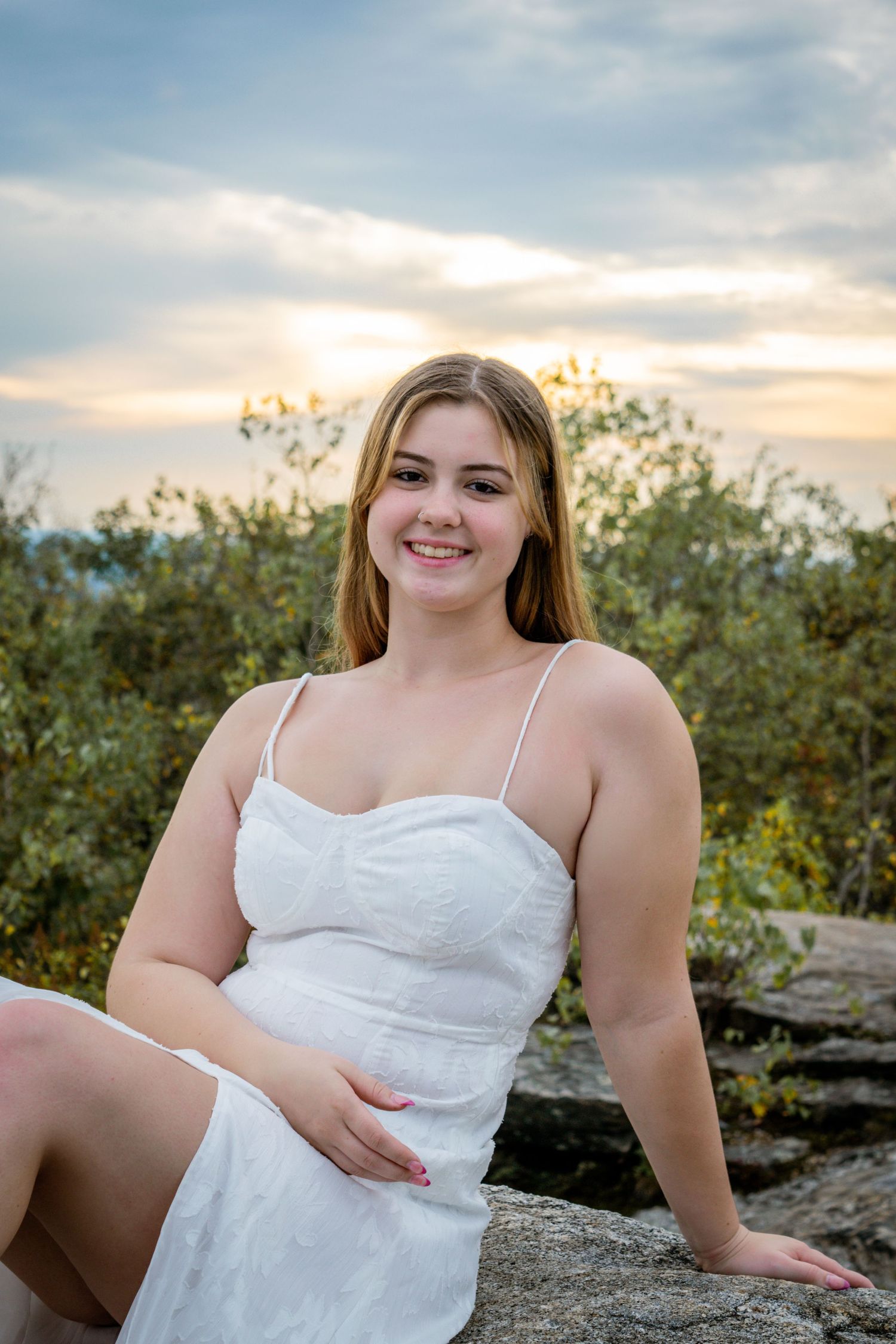 A person in a white sundress poses outdoors on rocks at sunset with greenery in the background.