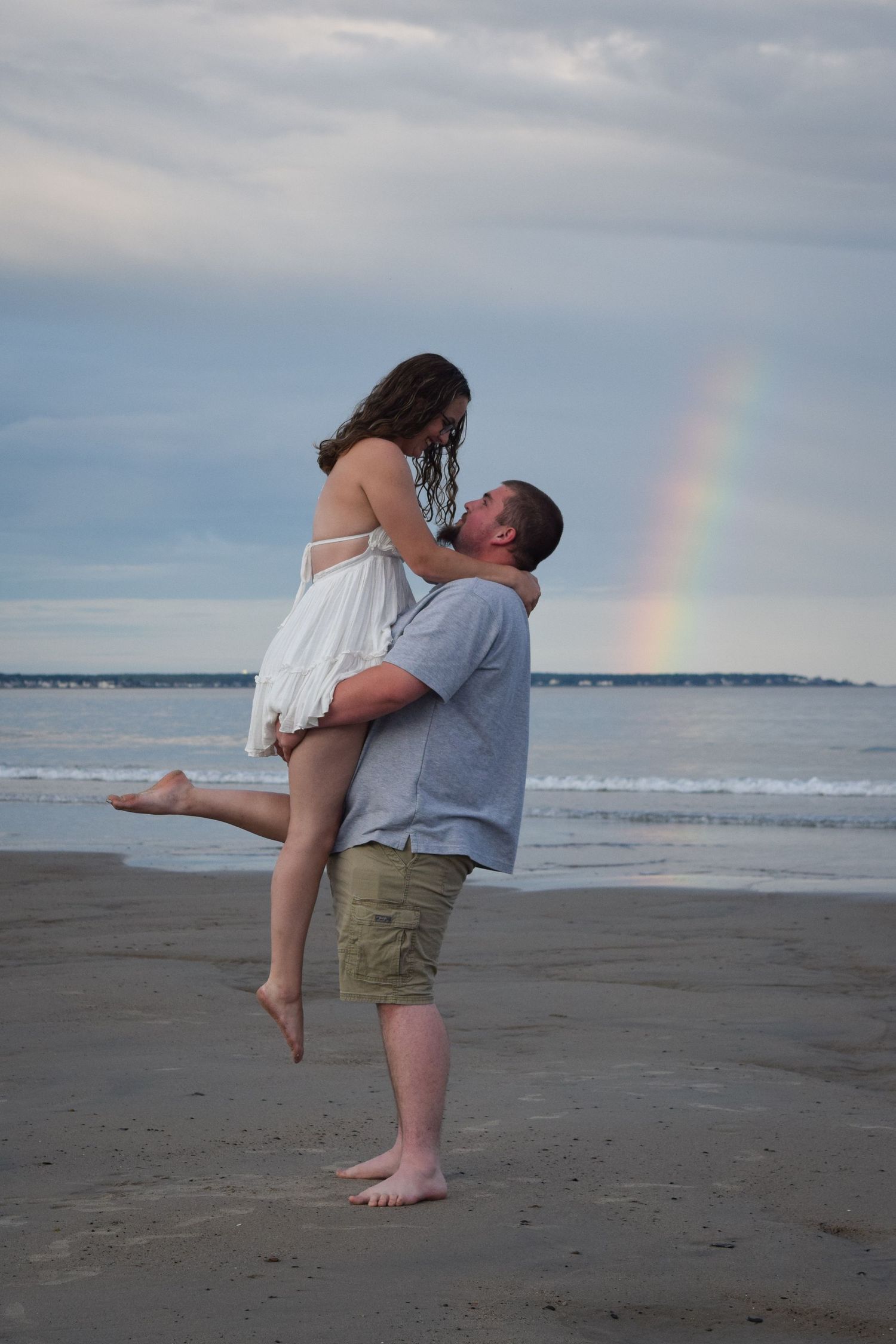 A couple embraces in a romantic kiss on a beach with a rainbow visible in the cloudy sky over the ocean.
