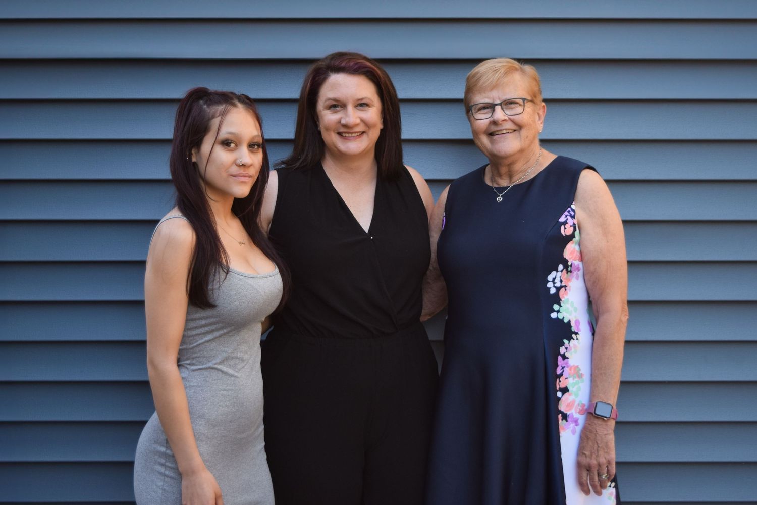 Three people stand together smiling in front of a blue siding wall on a summer evening.