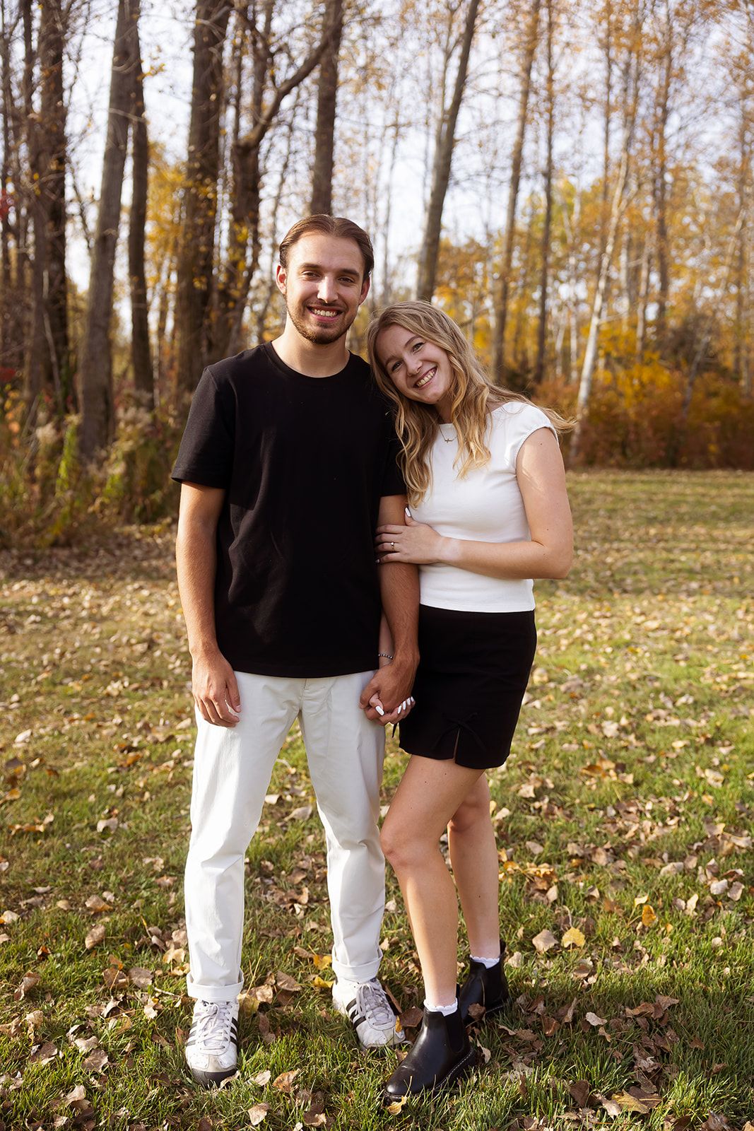 A couple stands together on fallen leaves in an autumn forest setting.