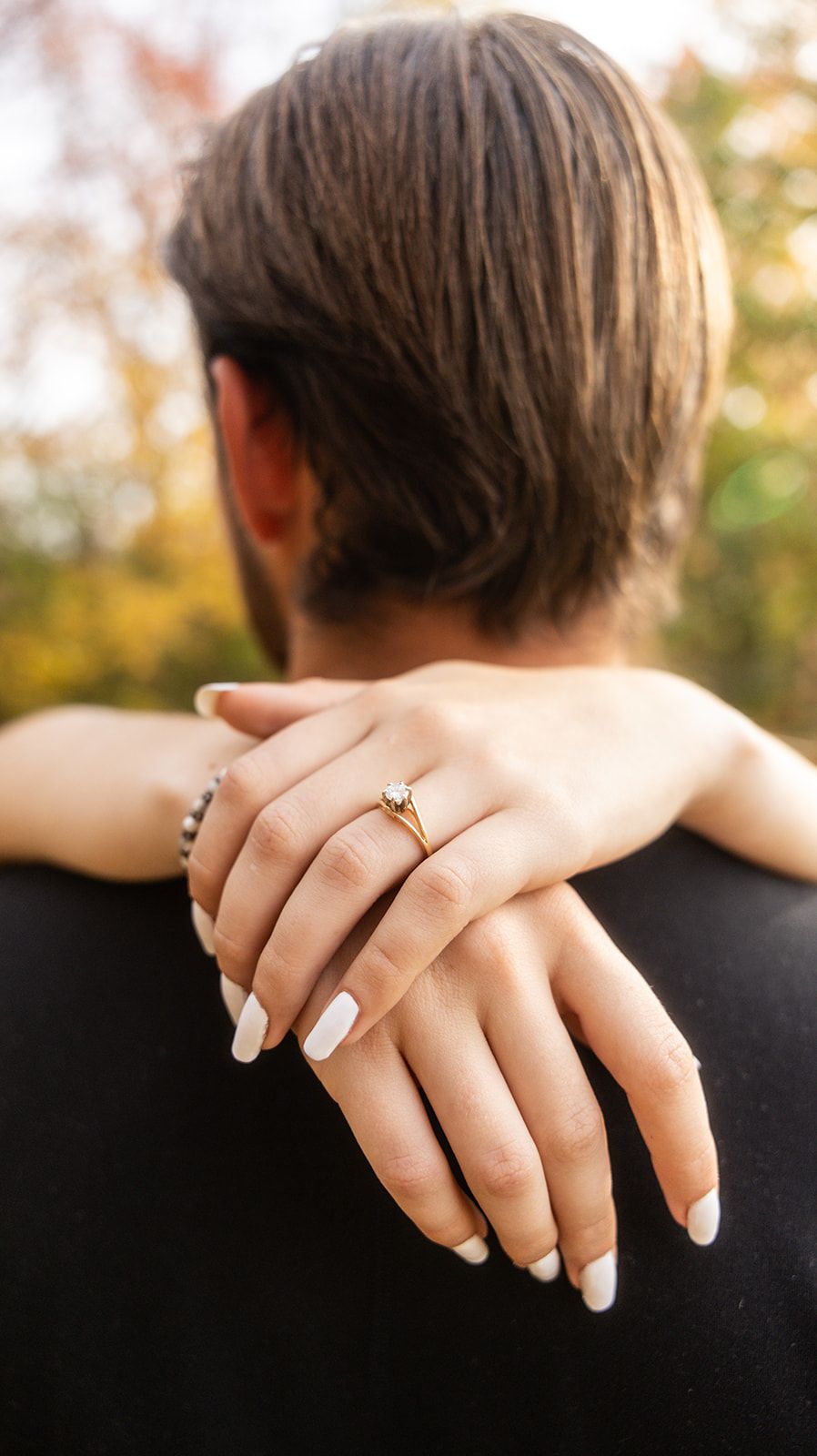 Close-up of hands showing an engagement ring against a blurred autumn background.