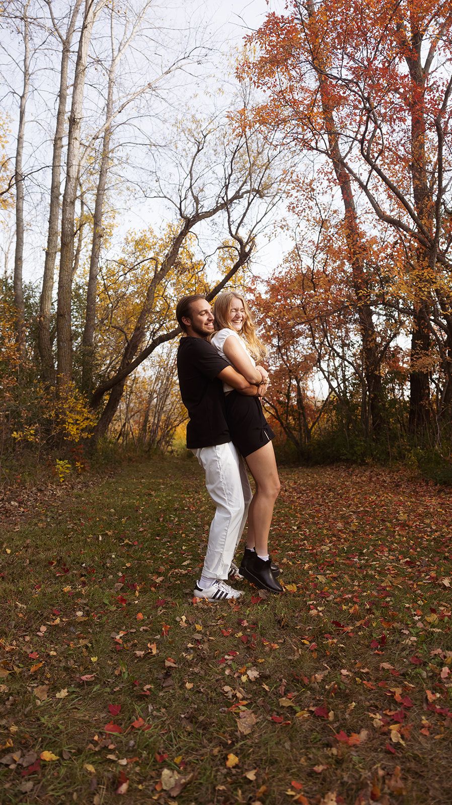 Playful couple in black and white outfits spinning in fall forest.