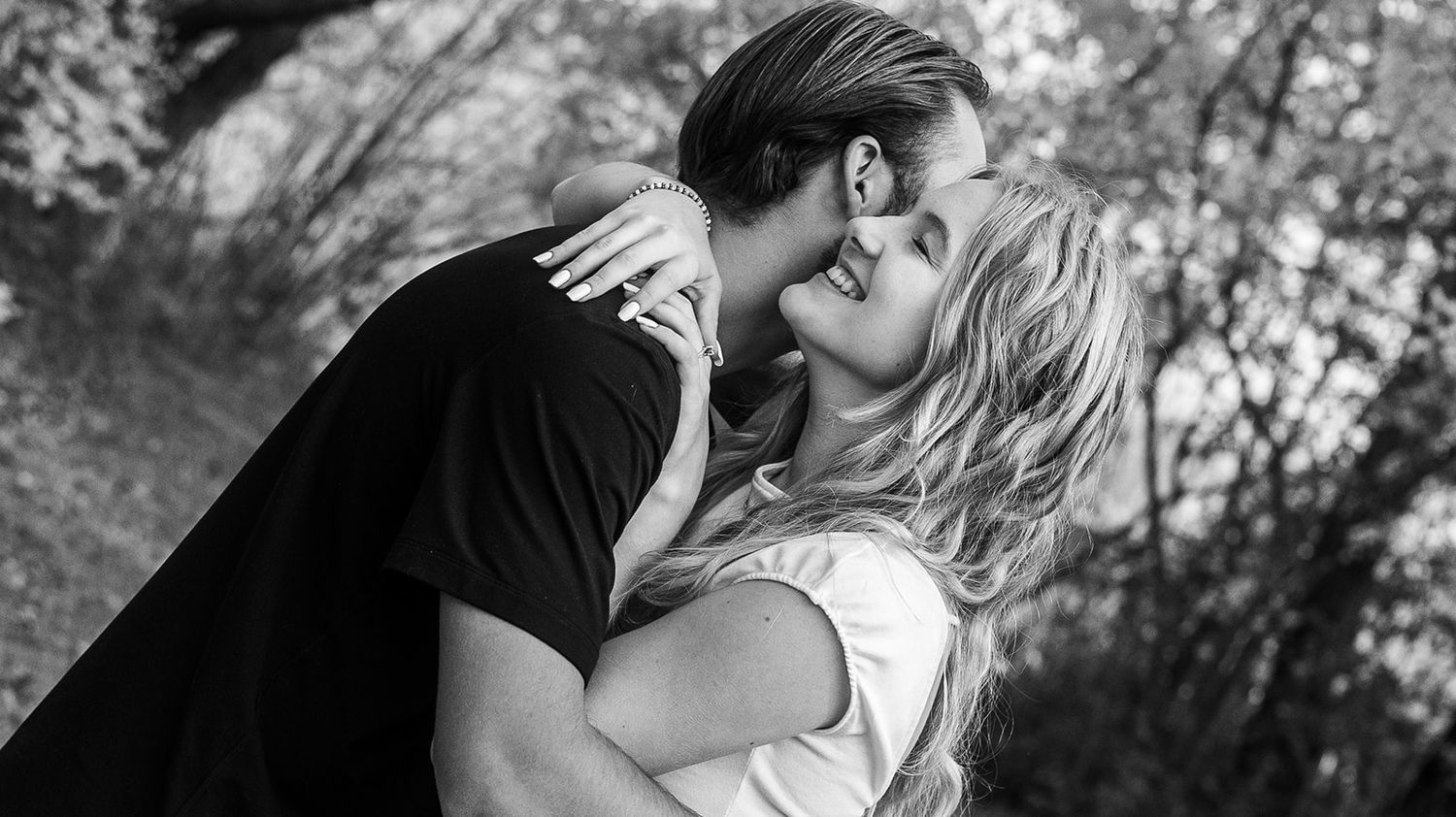A romantic couple shares an intimate moment in a wooded area with autumn foliage.