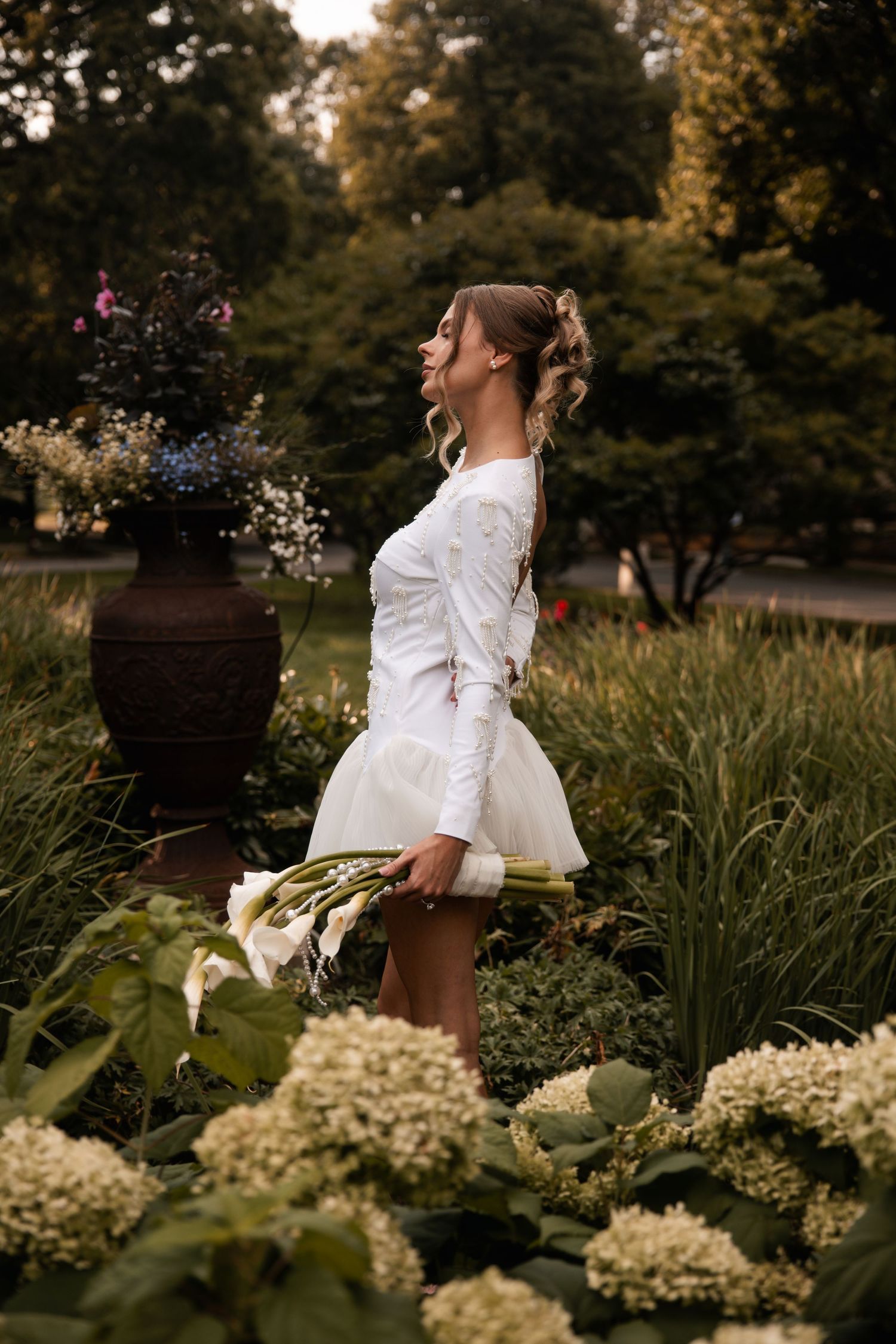 A side profile shot of a woman in white standing among blooming hydrangeas at dusk.