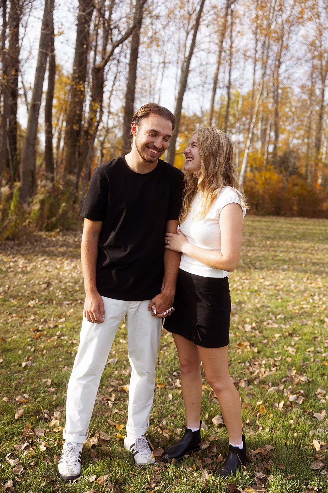 A couple stands together on fallen leaves in an autumn forest setting.