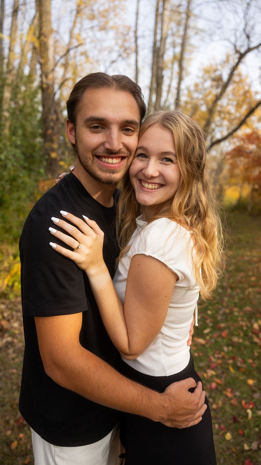 Couple embracing and showing off engagement ring in a wooded autumn setting.
