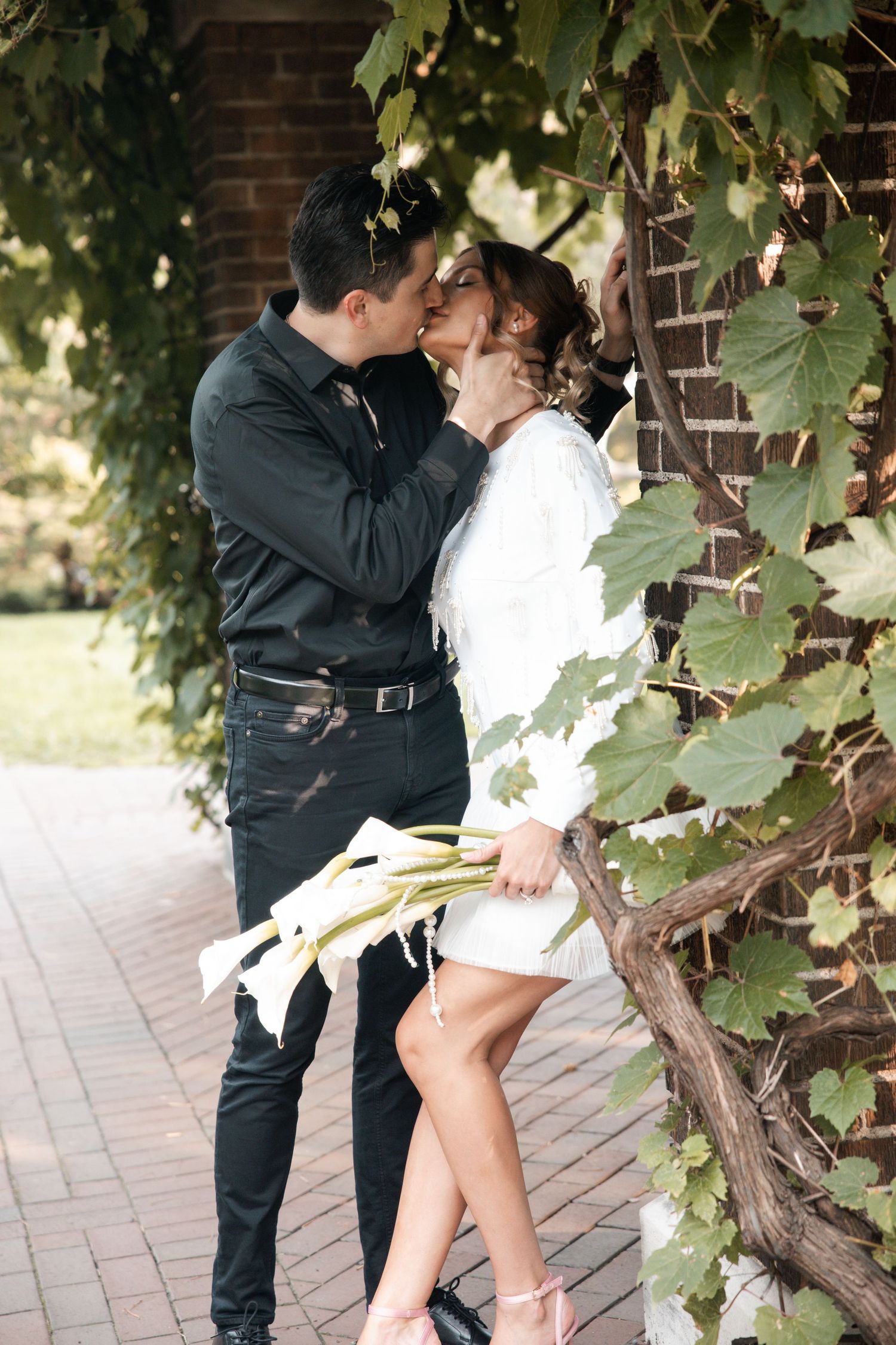 A romantic couple embraces near a vine-covered brick wall in an outdoor setting.