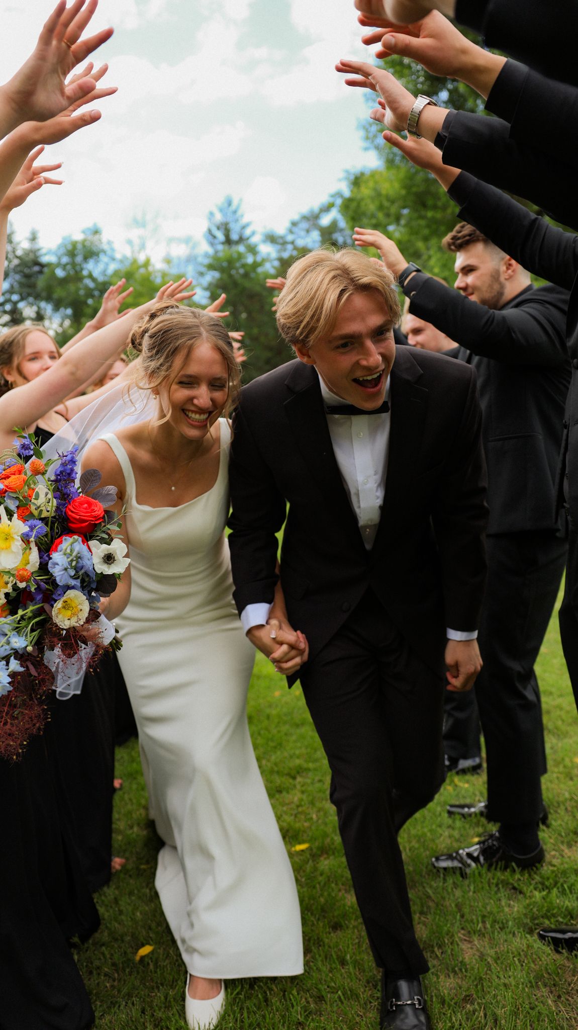 Wedding party celebrates with raised arms and laughter on a grassy lawn.