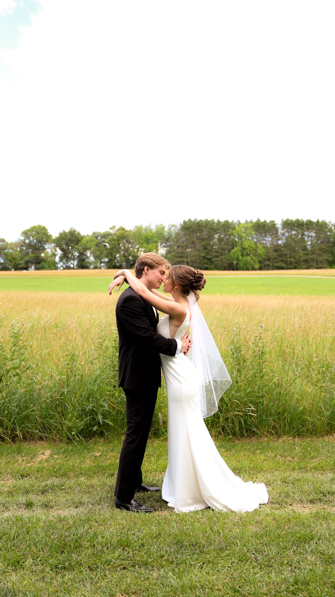 Wedding couple poses playfully in sunny meadow with trees in background.