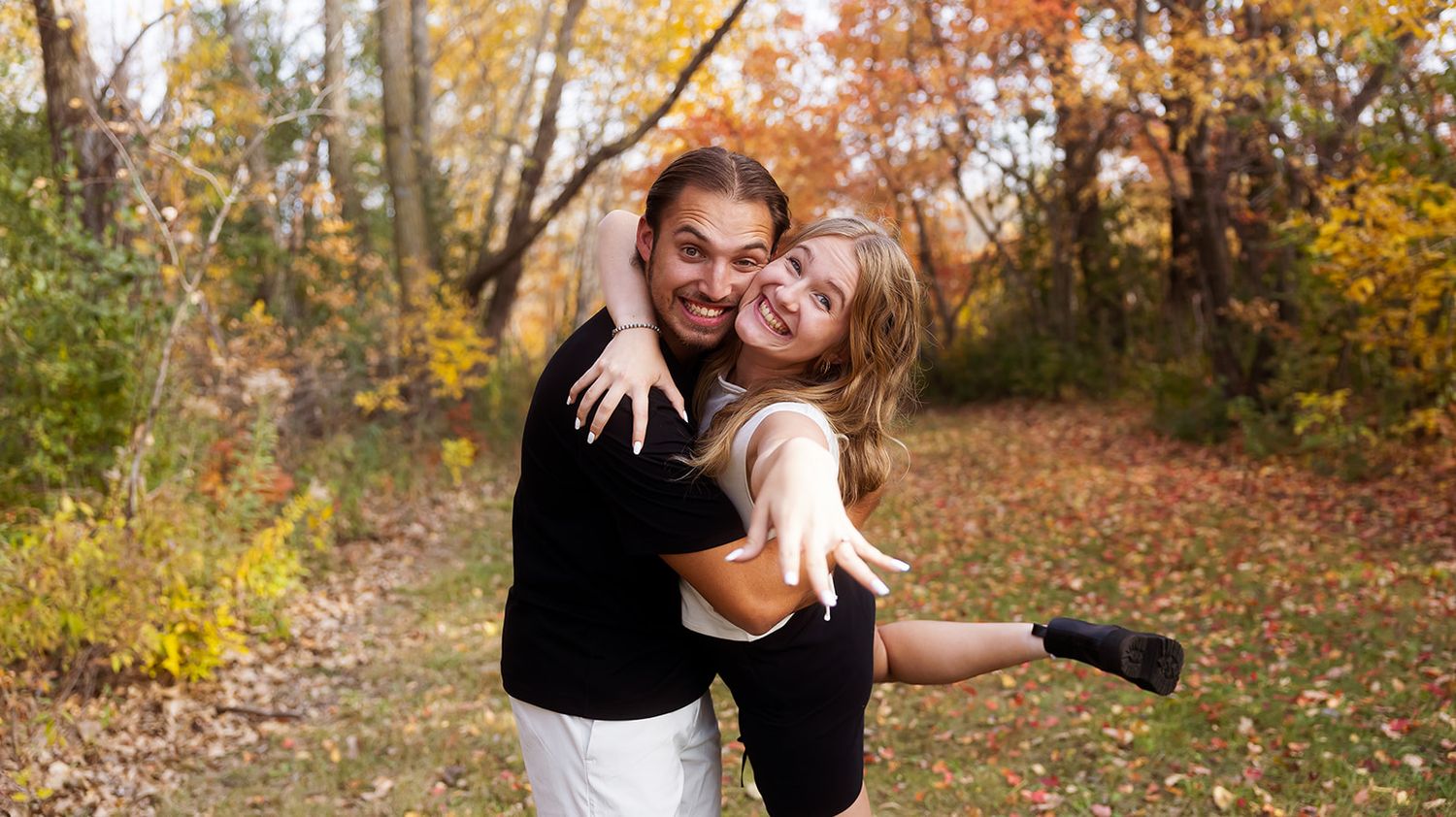 A couple embraces and laughs together in a forest during fall with golden leaves in the background.