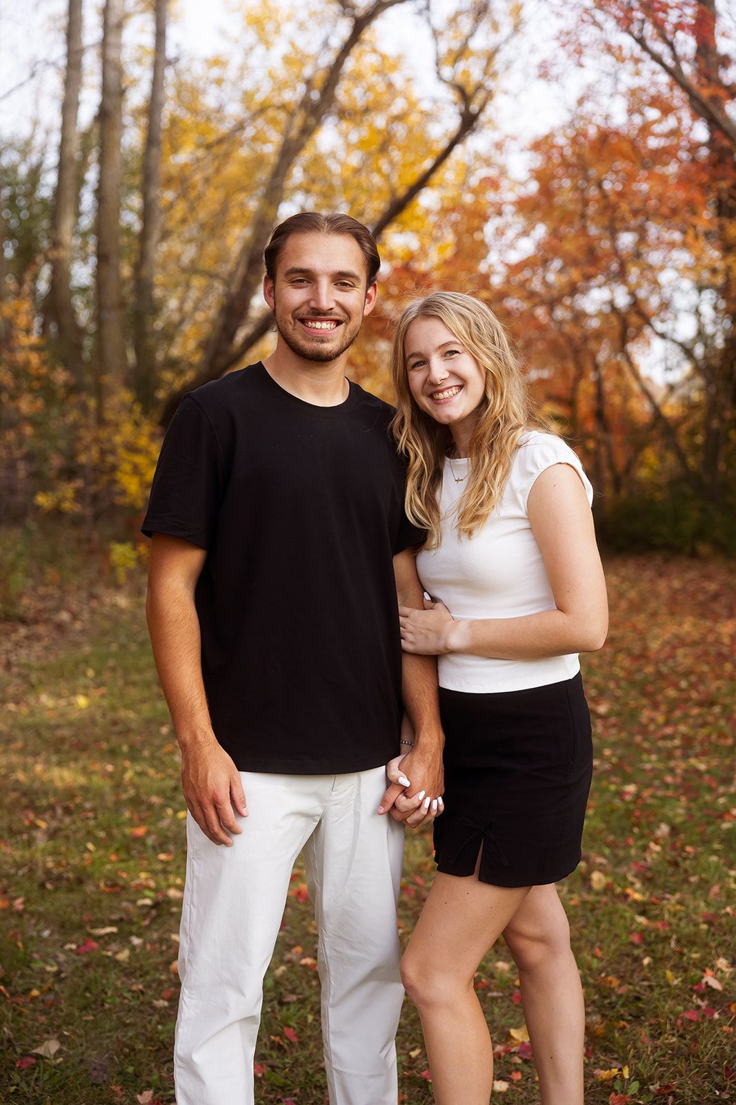 A couple poses together against vibrant fall foliage in a forest setting.
