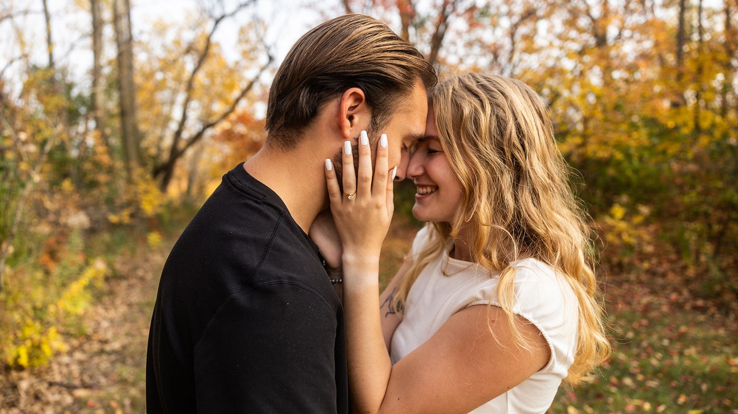 A couple embraces in an autumn forest setting with golden leaves visible.