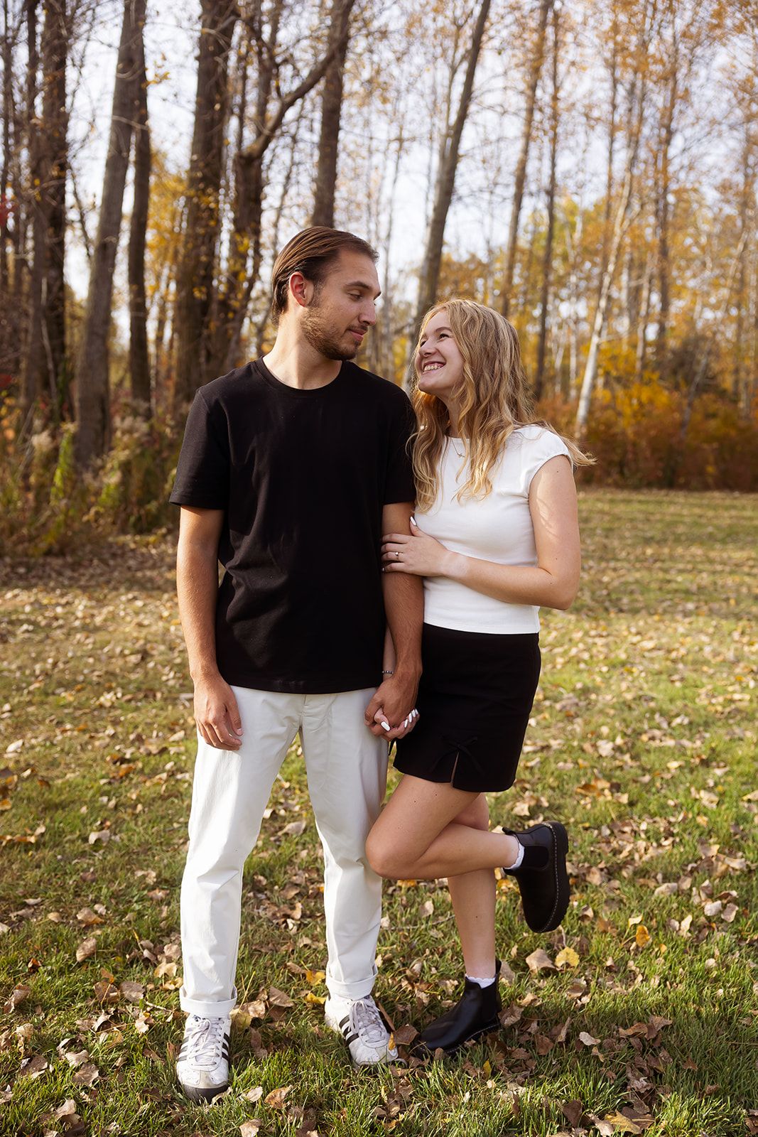 A couple poses casually together among autumn trees and fallen leaves.