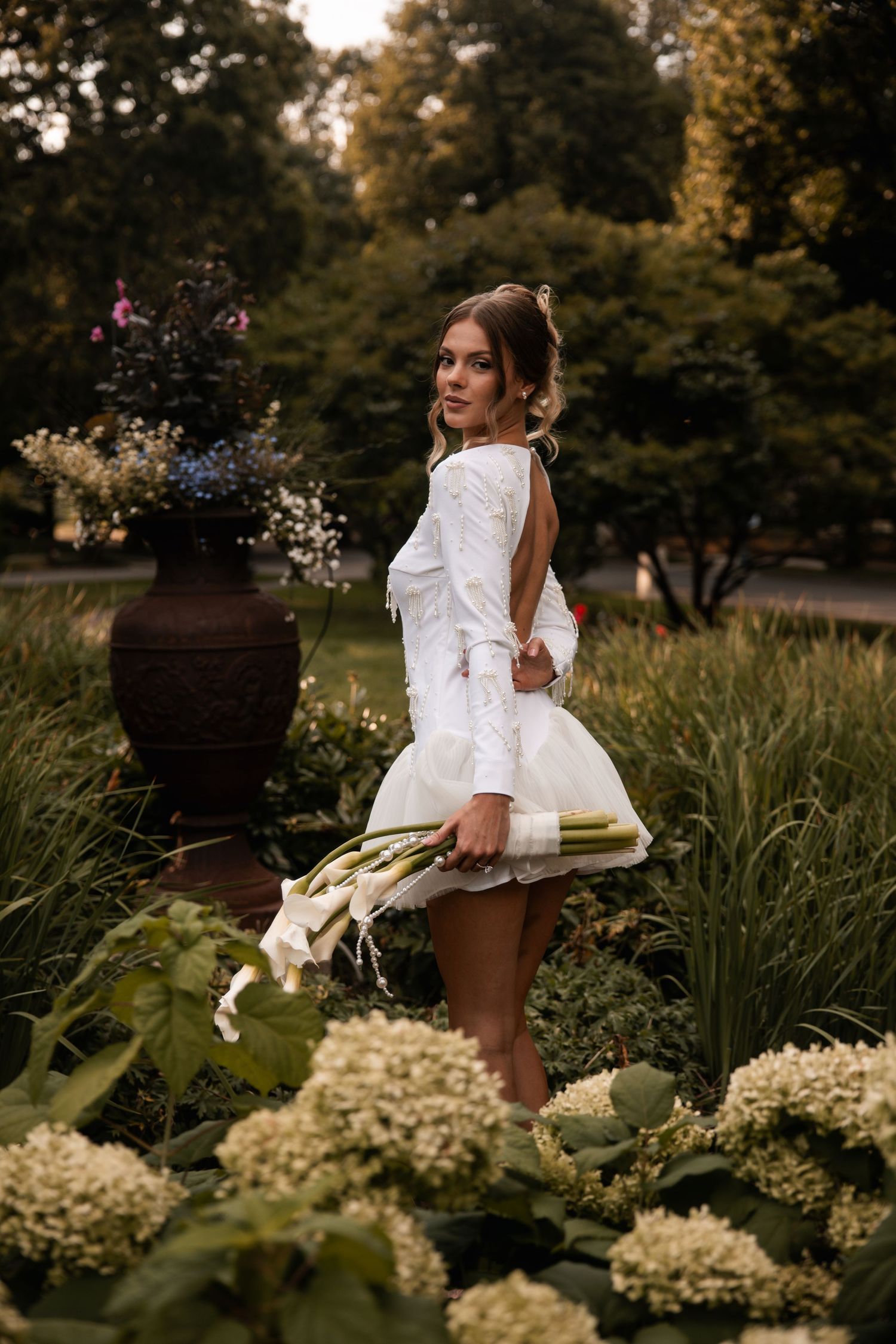 A woman in white stands gracefully among hydrangea bushes near a decorative garden urn.