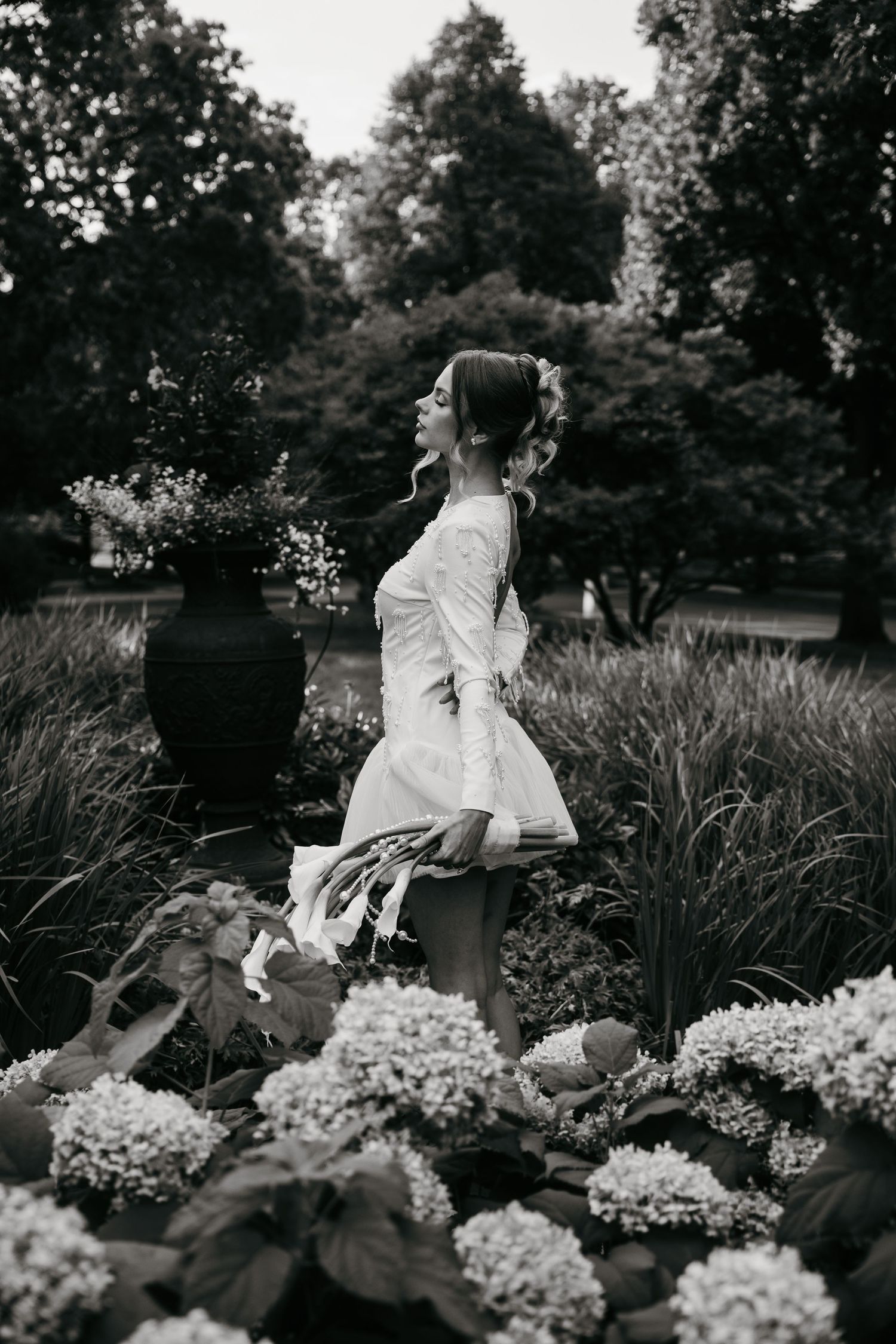 A bride in a short white dress stands among blooming hydrangeas in a garden setting.