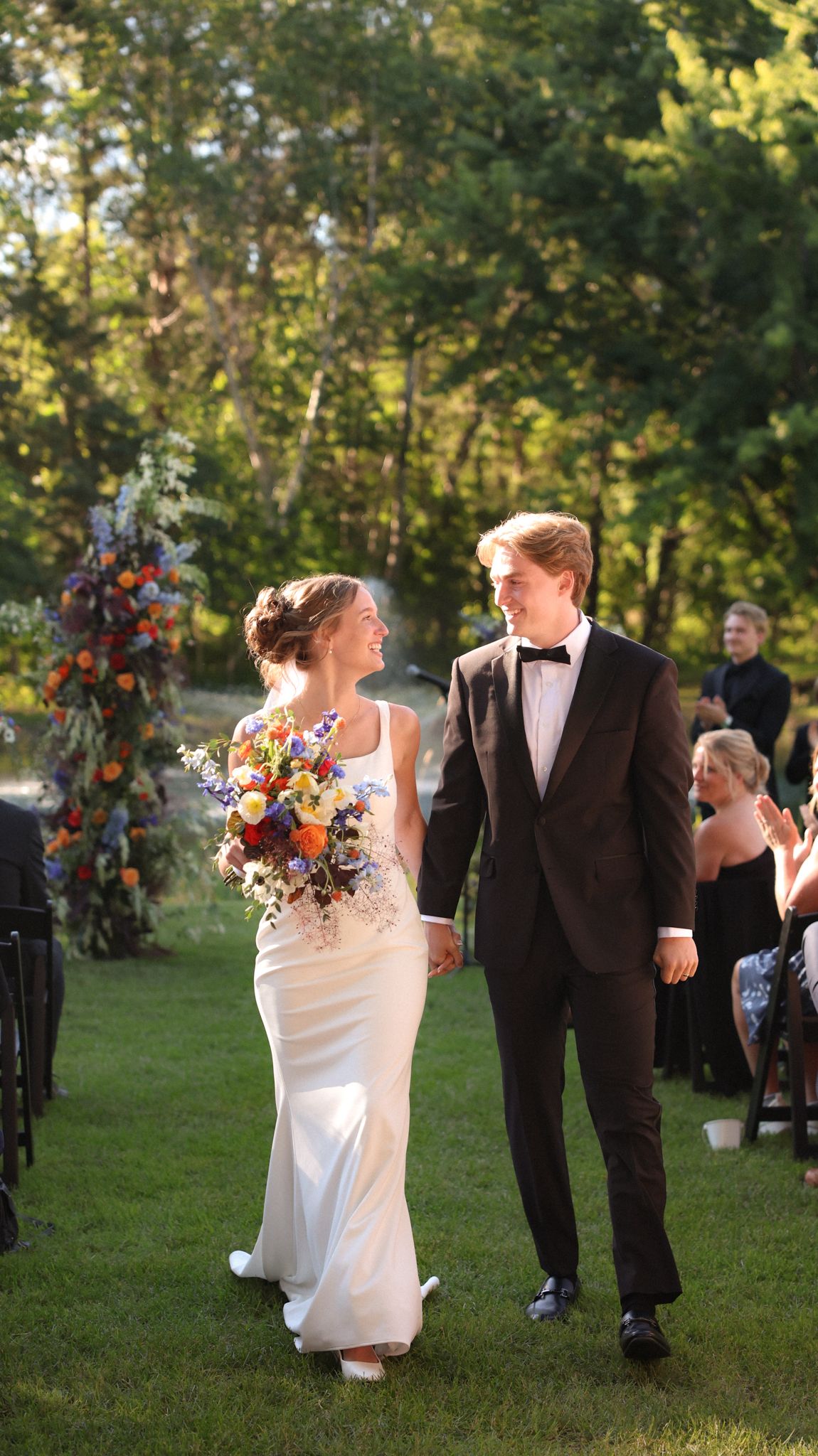 Wedding couple walks joyfully across a sunlit lawn during their outdoor ceremony.