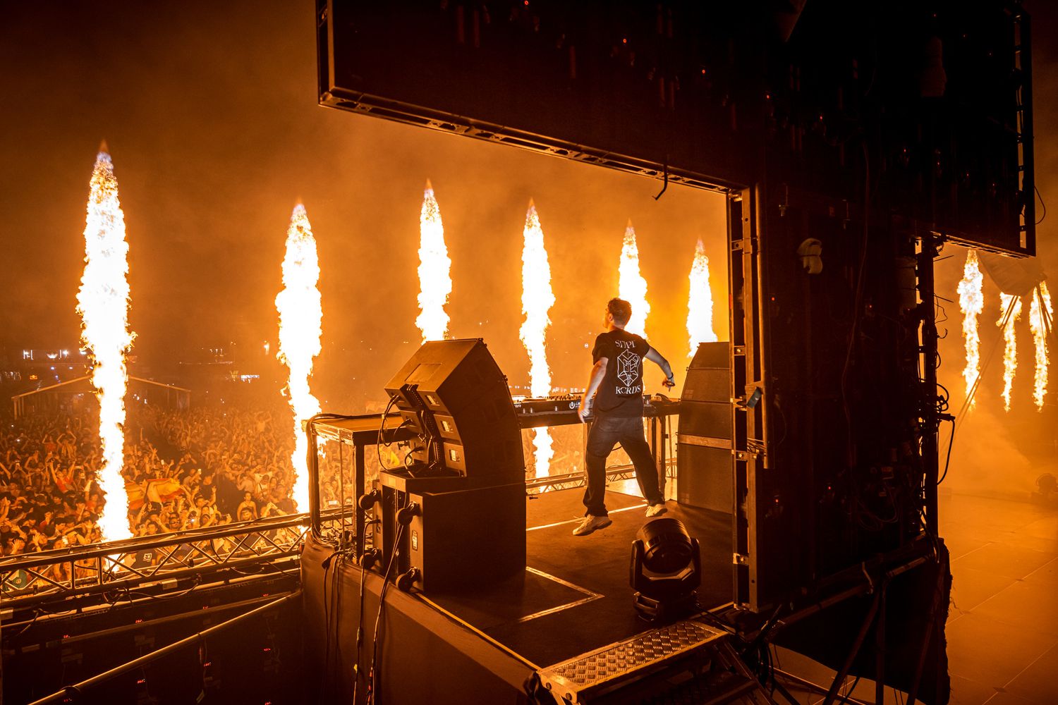 DJ performing on stage with dramatic fire pyrotechnics and large crowd at an outdoor music festival.