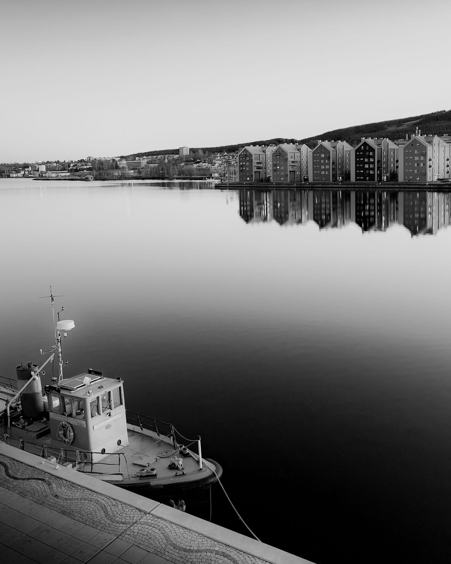 Black and white panoramic view of boats moored at a tranquil harbor with buildings in the background.