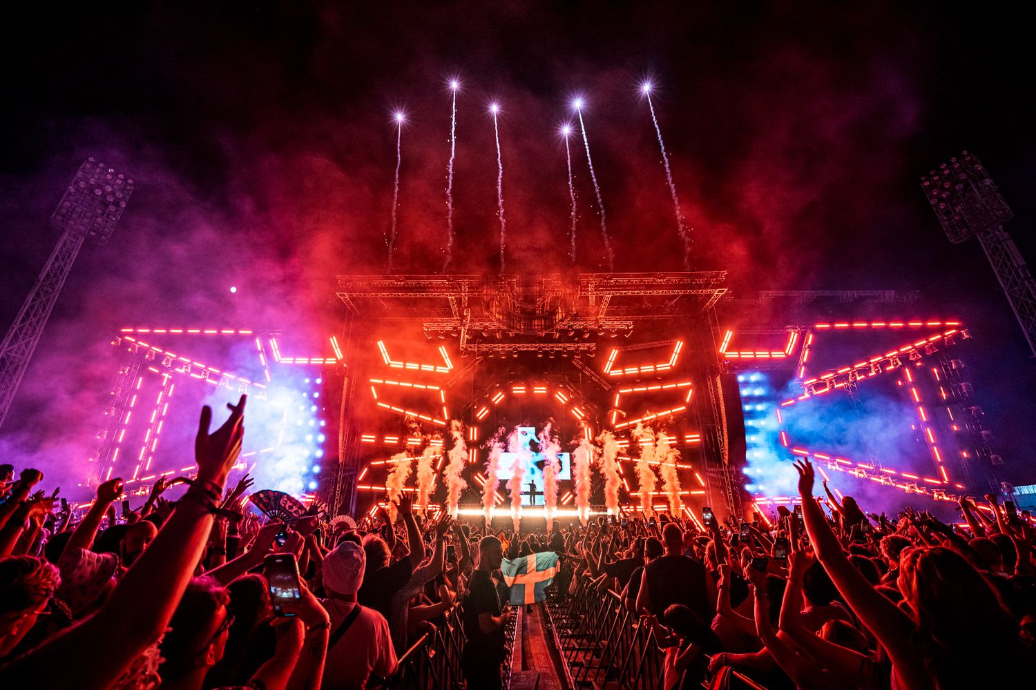 Massive outdoor music festival stage with red neon lights, fireworks, and crowd cheering under a vibrant night sky.
