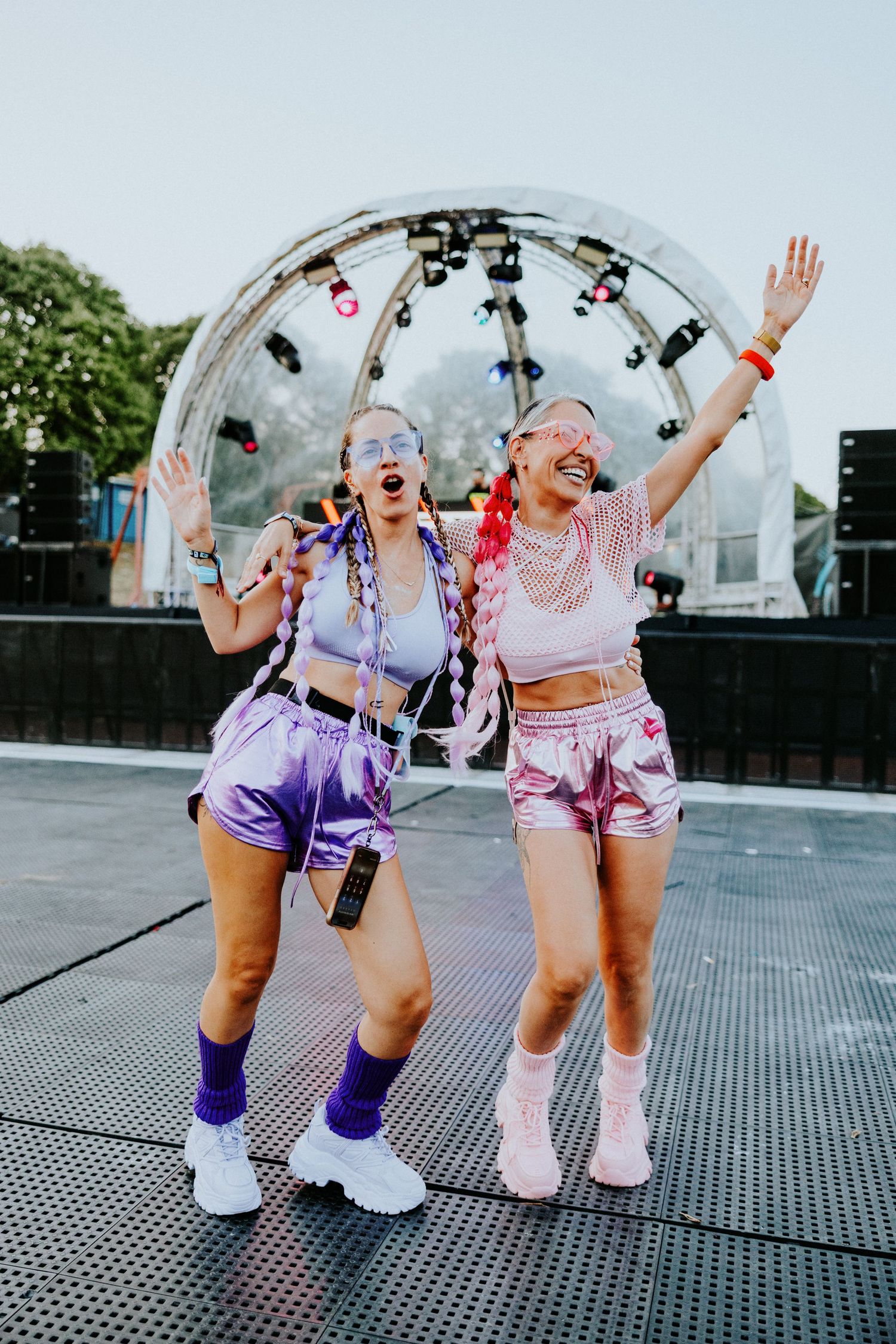 Two women in colorful festival outfits dancing joyfully in front of a stage with lights.