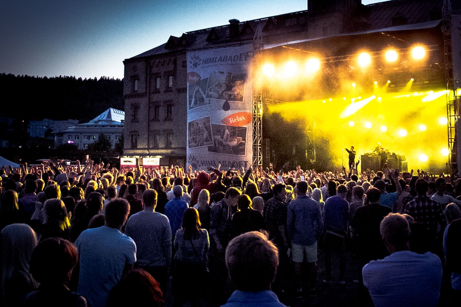 Large outdoor concert crowd gathered at night with bright yellow stage lights illuminating the scene.