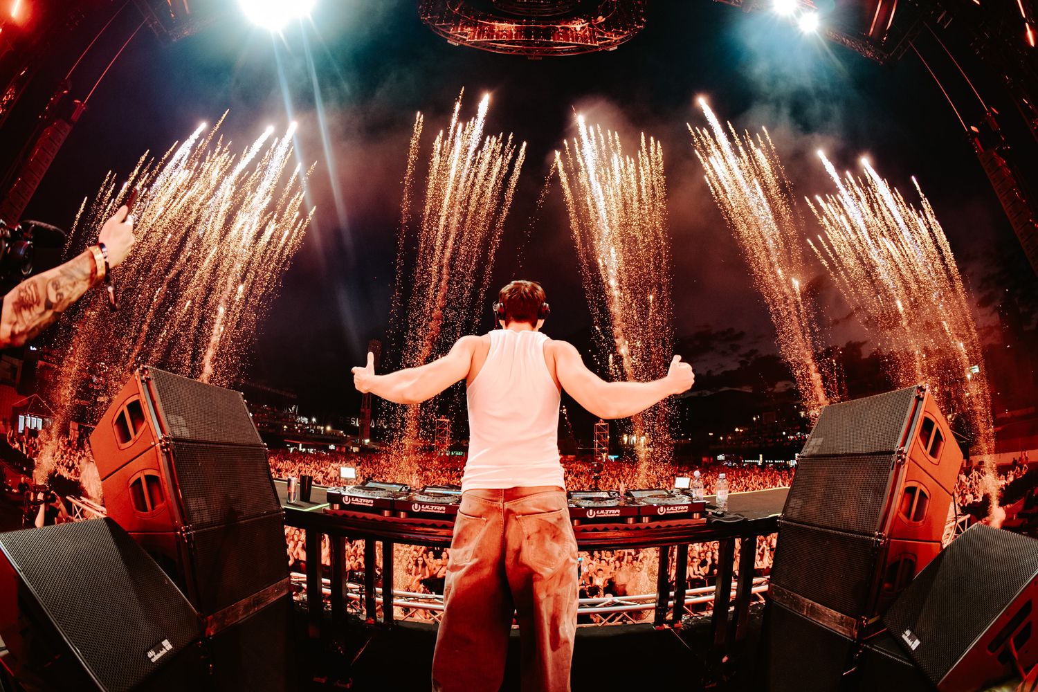 DJ performs on stage with arms raised as golden fireworks explode behind him at a massive music festival.