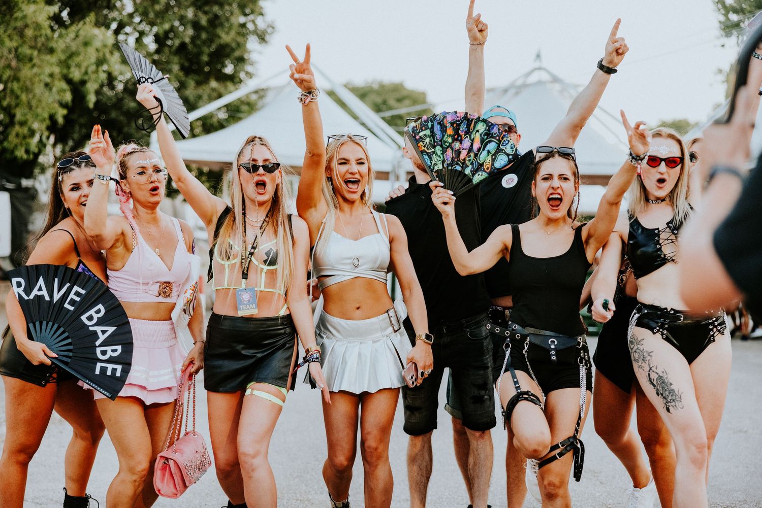 Group of women in festival outfits celebrating at a rave, one holding a 'Rave Babe' sign.