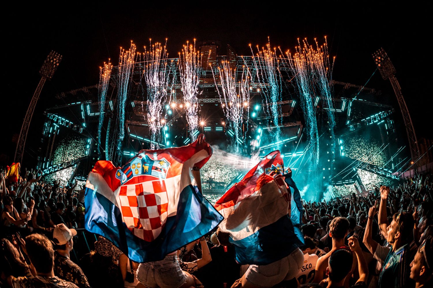 Crowd waving Croatian flags at a music festival with fireworks and stage lights illuminating the night sky.
