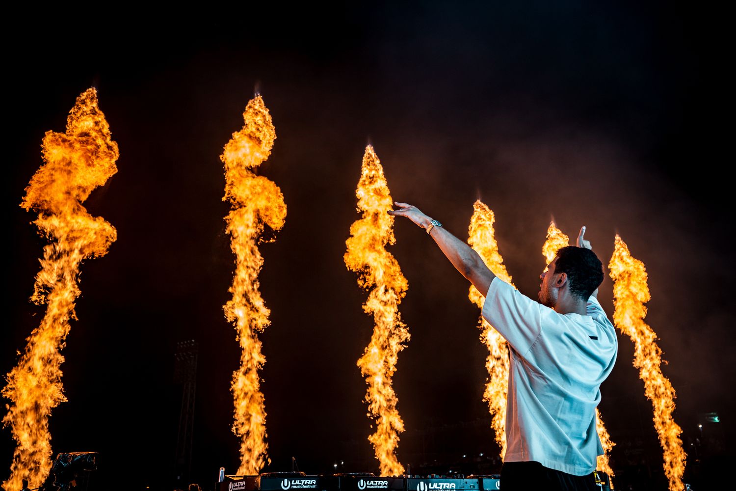 DJ performing on stage with dramatic columns of fire pyrotechnics blazing behind him at a night concert.