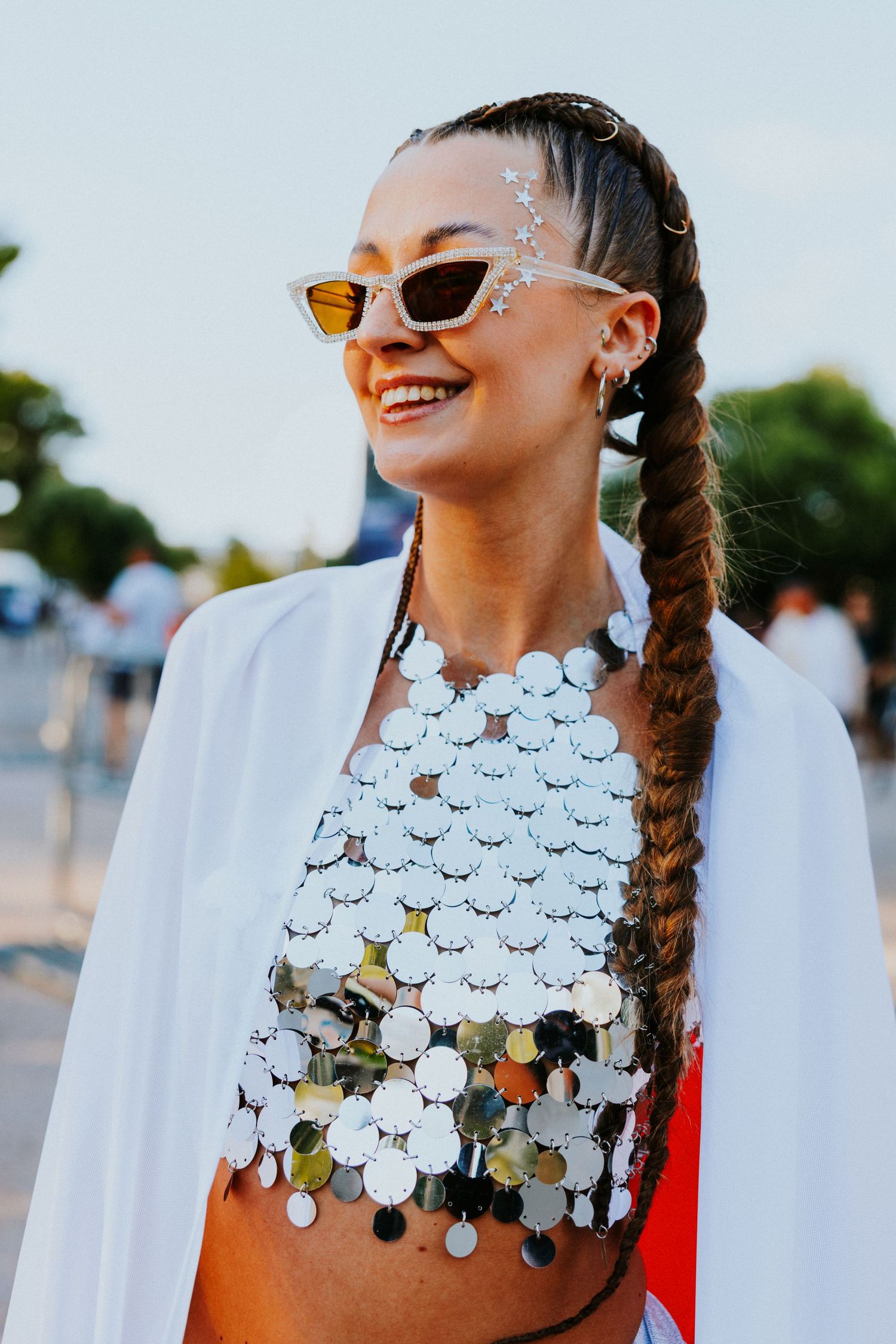 Smiling woman at festival wearing silver coin crop top, white jacket, braided hair, small sunglasses, and glitter face gems.