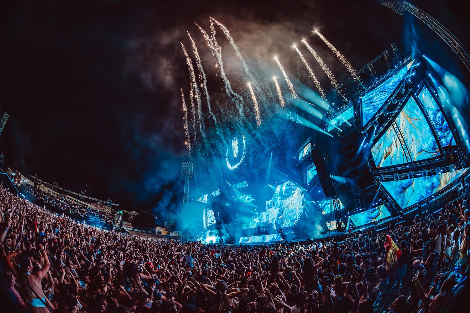 Massive outdoor music festival with stunning blue stage lights, fireworks, and enormous crowd at night.