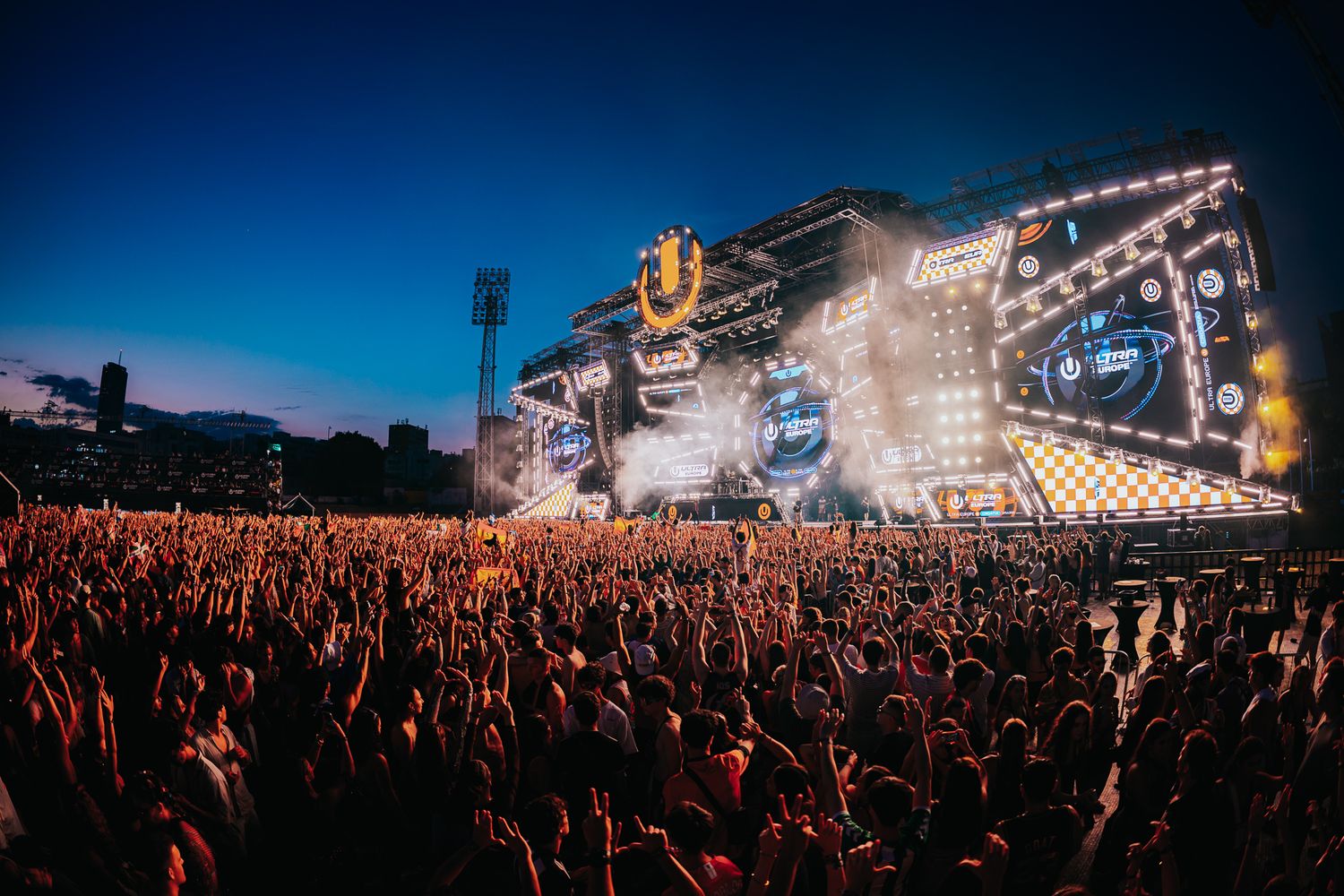 Massive crowd at an outdoor music festival with a spectacular illuminated stage and dramatic twilight sky.